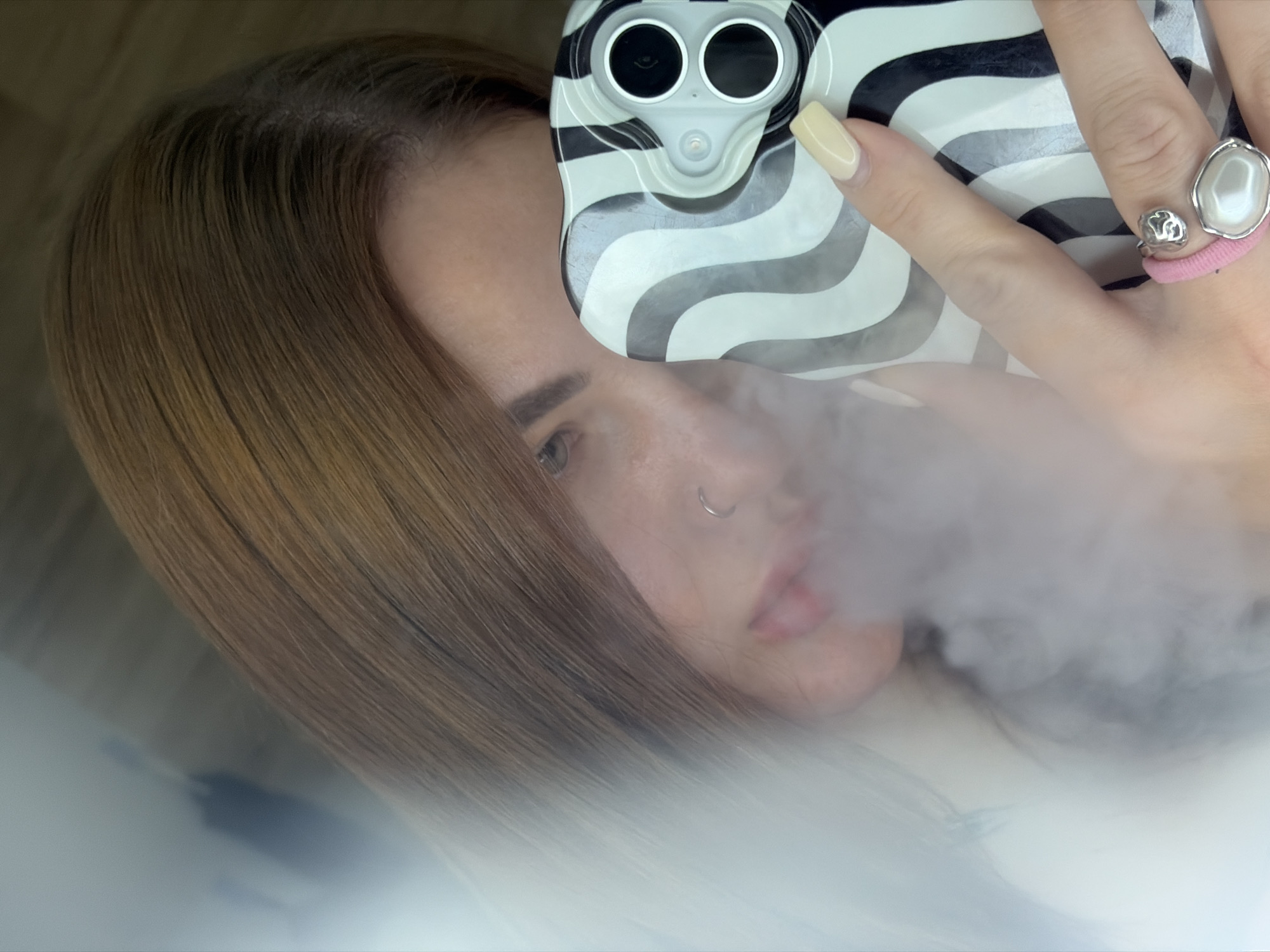 A woman with long, brown hair and a nose ring is taking a selfie with a smartphone. The phone has a black and white wavy striped case. She is holding the phone with her right hand, and the ring finger has a pink hair tie with two silver rings on top. A cloud of vapor is present near her mouth. The photograph was likely taken in a personal space, but no specific details indicate it is in Krasnodar, Russia.
