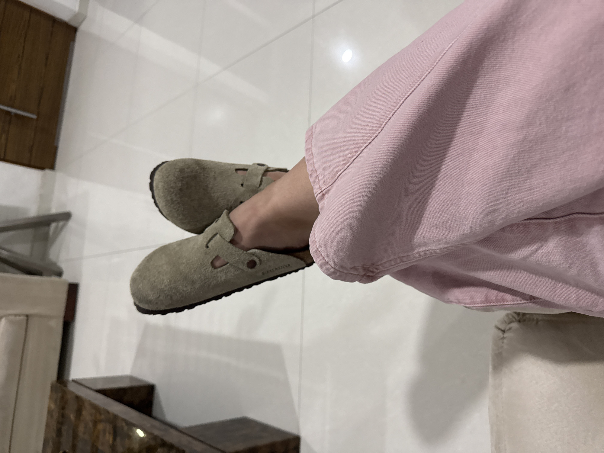 The image shows a pair of feet wearing olive green suede Birkenstock Boston clogs. The person is wearing pink denim pants. The background consists of white tiled flooring and a piece of dark wood furniture. The location is not possible to determine based on the given image.