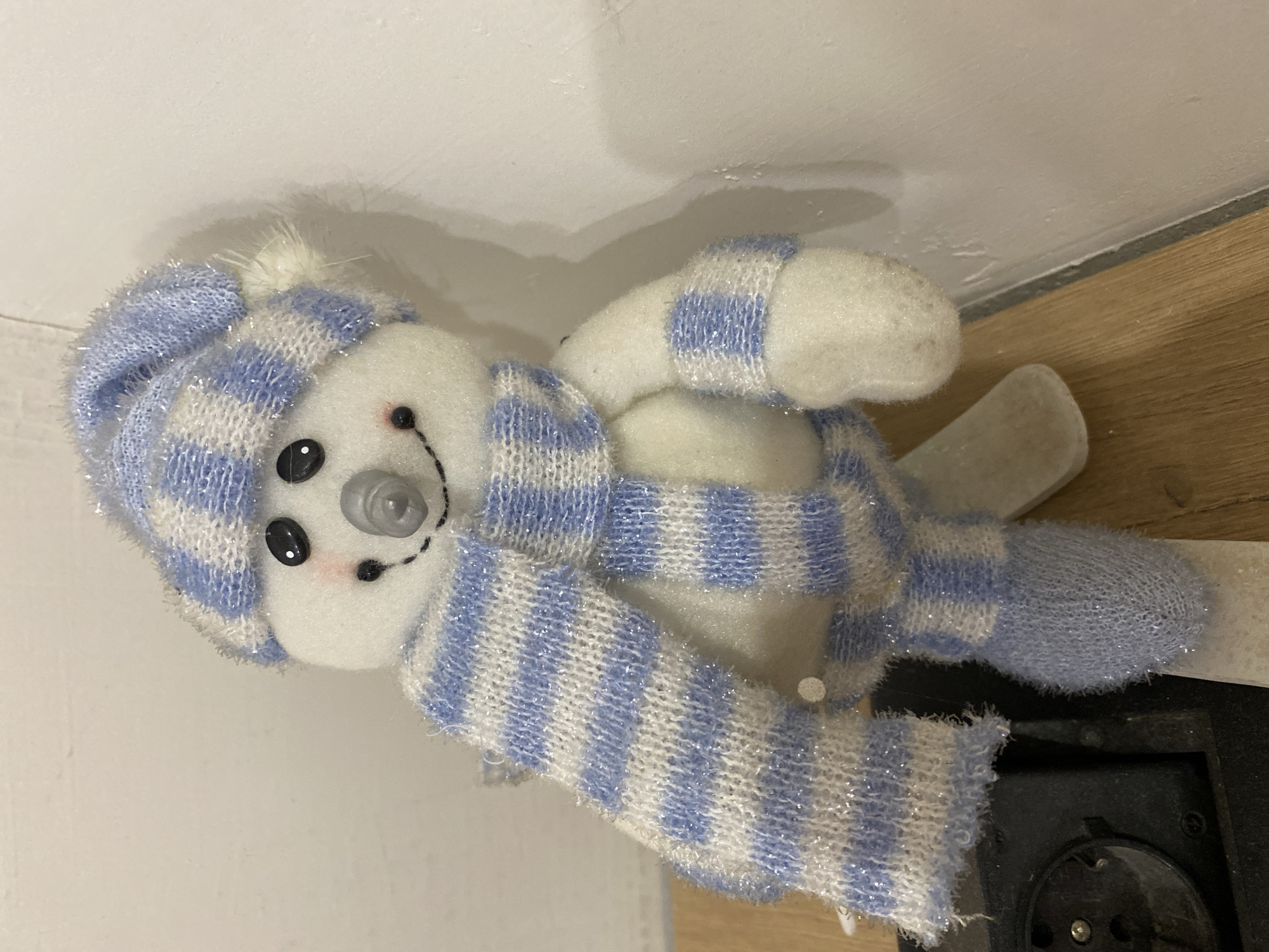 A decorative snowman figurine is depicted standing upright in what appears to be an interior setting in Chaikovsky, Russia. The snowman features a white, plush body with black spherical eyes, a small grey conical nose, and a dotted black smile with subtle red cheek accents. It is dressed in a light blue and white striped knitted beanie with a white pom-pom on its right side, and a matching striped scarf wrapped around its neck. The knitted material contains shimmering silver threads. Its lower arms and feet are also covered in similar blue and white striped or solid blue knitted fabric. The figurine is positioned on a light brown wooden surface, possibly a shelf or floor, and leans against a plain, off-white wall. In the lower-left foreground, a portion of a black electrical socket or similar circular component is visible on the wooden surface.