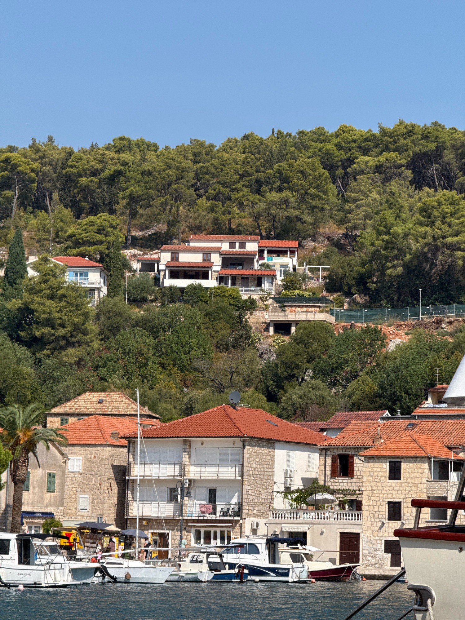 This image depicts a waterside view of Jelsa, Croatia, showcasing a row of stone buildings with red tile roofs lining the harbor. Several boats, including sailboats and motorboats, are docked in the calm blue water. In the background, a lush green hillside covered in trees rises behind the buildings, with more houses visible nestled amongst the foliage. The sky is clear and blue. There are no discernible people in action or interaction.