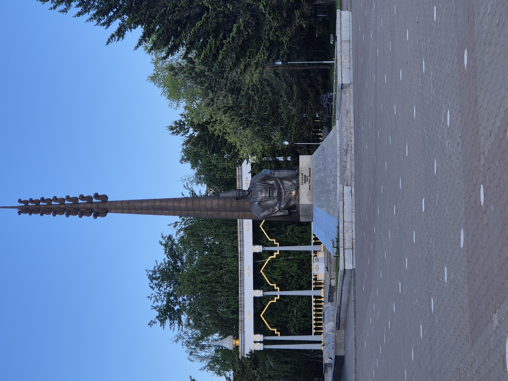 The image shows the Abai Qunanbaiuly monument in Almaty, Kazakhstan. The monument consists of a bronze statue of the poet Abai seated on a stone pedestal, behind which rises a tall, stylized column topped with several abstract figures. The monument is surrounded by a decorative fence with white pillars and gold accents. In the background, lush green trees fill the scene. The foreground is a paved area with white markings. The sky is clear and blue.
