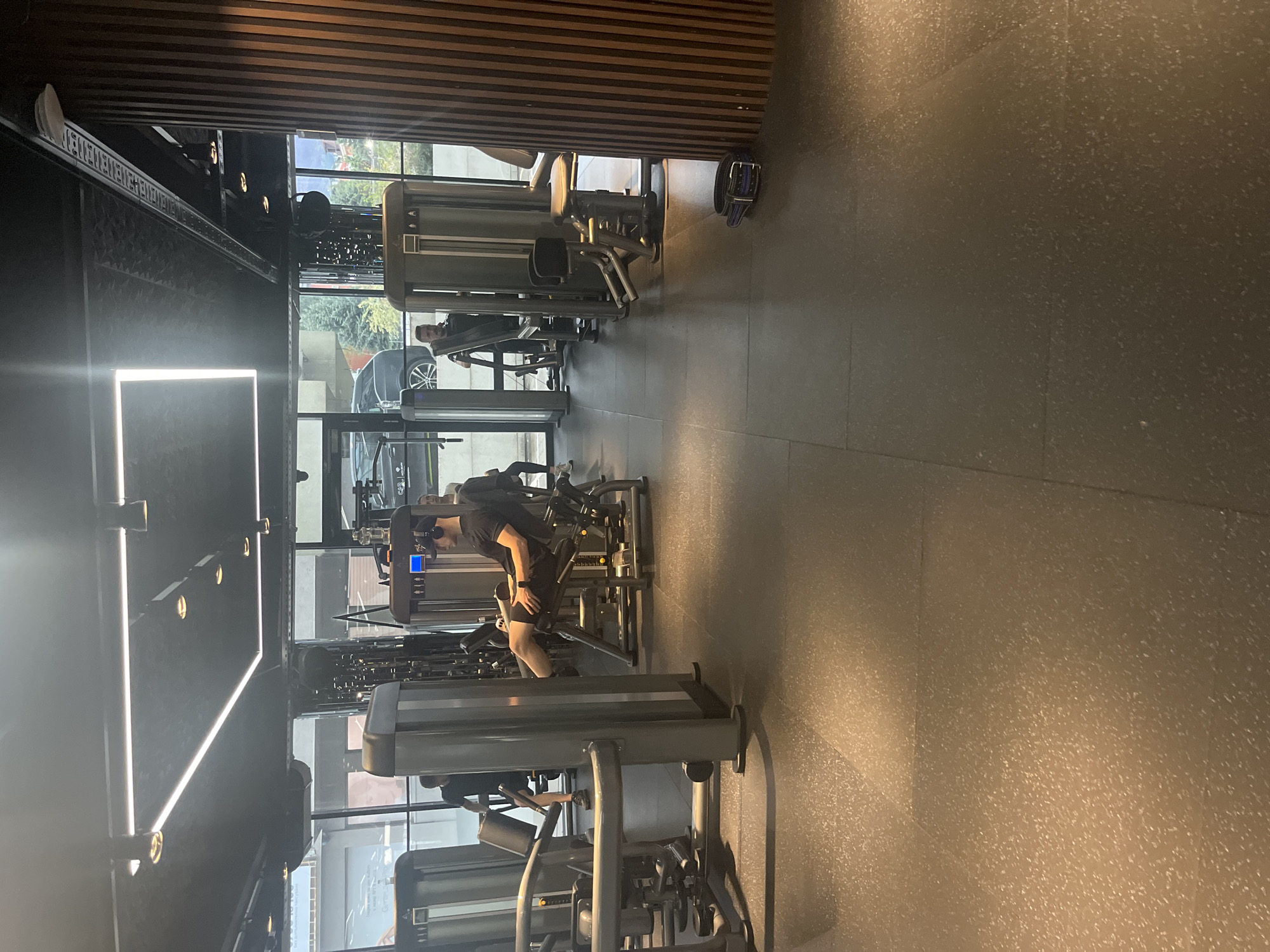 The image depicts the interior of a modern gym or fitness center in Sofia, Bulgaria, oriented to be viewed landscape. Natural light streams in through large floor-to-ceiling windows on the left side, illuminating the dark tiled floor and various gym equipment.
In the foreground, a person wearing a dark t-shirt and shorts, possibly with headphones, is actively using a leg curl or similar selectorized machine. Further back, another individual is visible, also engaged with gym equipment near the windows. The gym features numerous dark-colored exercise machines, arranged in rows.
Notable architectural details include a ceiling and upper wall section covered in dark wooden slats, adding a warm, contemporary accent. The lighting design incorporates both linear LED light strips running along a wall and ceiling, and several recessed spotlights, creating bright, even illumination. Through the windows, parts of a parked car and some greenery are visible, indicating an urban setting.
On the dark tiled floor, near one of the machines, a black and blue dog collar is distinctly visible. High on a white beam stretching across the upper part of the room, the text "STRENGTH. CONDITIONING. PERFORMANCE." is clearly legible. The overall impression is one of a well-maintained, functional, and aesthetically pleasing fitness environment during daylight hours.
