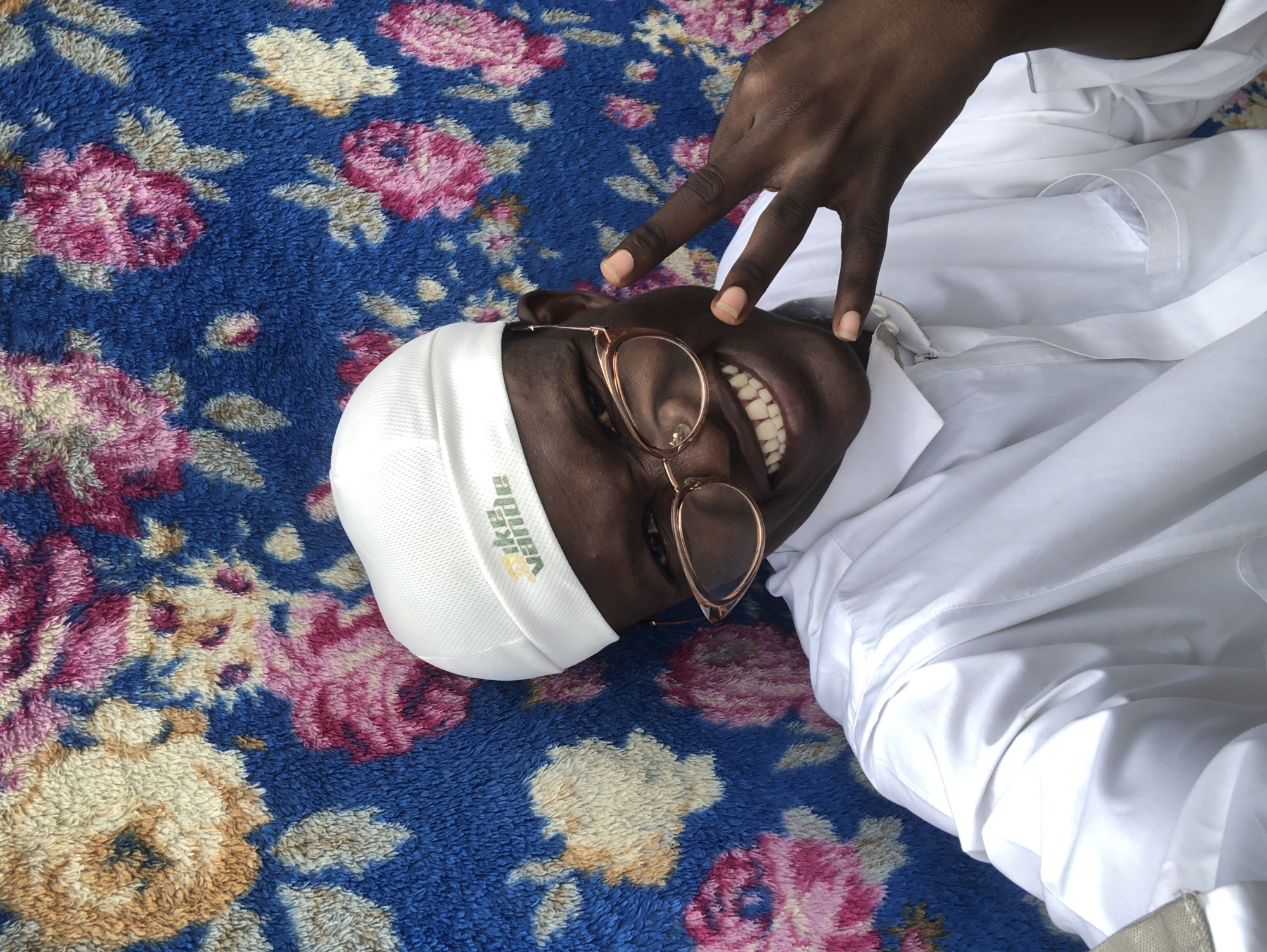In Ado, Nigeria, a young man with dark skin and a wide smile is captured in a selfie against a vibrant, floral-patterned backdrop. He is wearing a white kufi cap, spectacles with a rose-gold frame, and a white long-sleeved shirt, possibly a thobe. His right hand is raised, forming a peace sign with his index and middle fingers. The background is a textured fabric with a blue base color, adorned with an array of large, stylized flowers in shades of pink, yellow, and beige, creating a lively and somewhat busy visual setting.