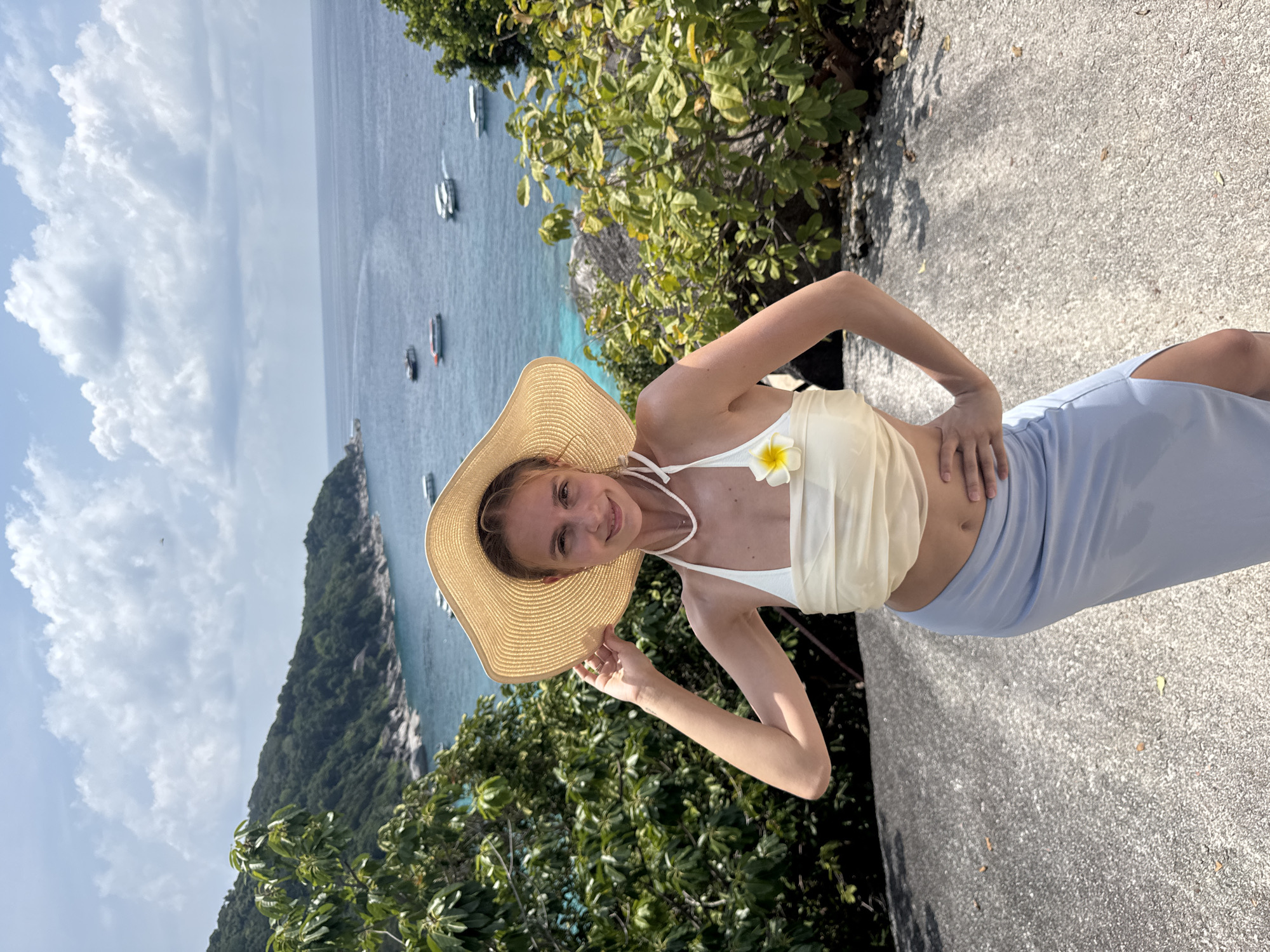 A woman stands on a paved surface near a lush green hillside and an ocean view in Lam Kaen, Thailand. She is wearing a straw hat, a white top with a flower, and a light blue skirt. She poses with one hand holding the brim of her hat and the other on her hip. The ocean is visible in the background, dotted with several boats.