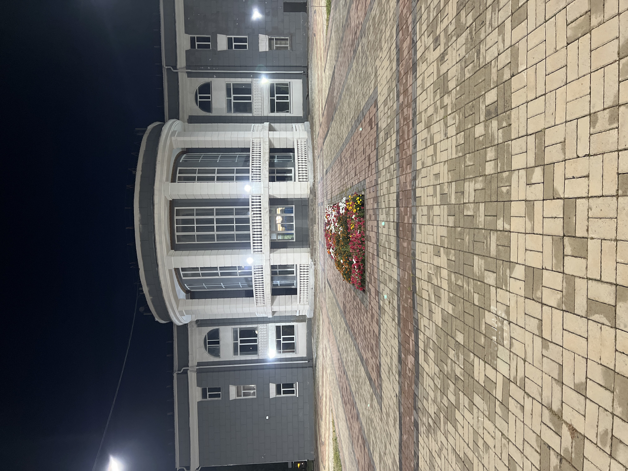 The image shows the exterior of a building at night, likely the city administration building in Qarağandı, Kazakhstan. The building is modern with a large, curved facade featuring numerous windows and white decorative columns. A balcony with white railings is visible in front of the central section. The building is illuminated by artificial lights. In front of the building, there is a flowerbed with a variety of colorful flowers, including red, pink, white, and yellow blooms. The foreground consists of a paved area with a pattern of rectangular bricks in shades of beige and reddish-brown. The sky above is dark, indicating it is nighttime. The scene appears to be an outdoor plaza or entryway to the administrative building.