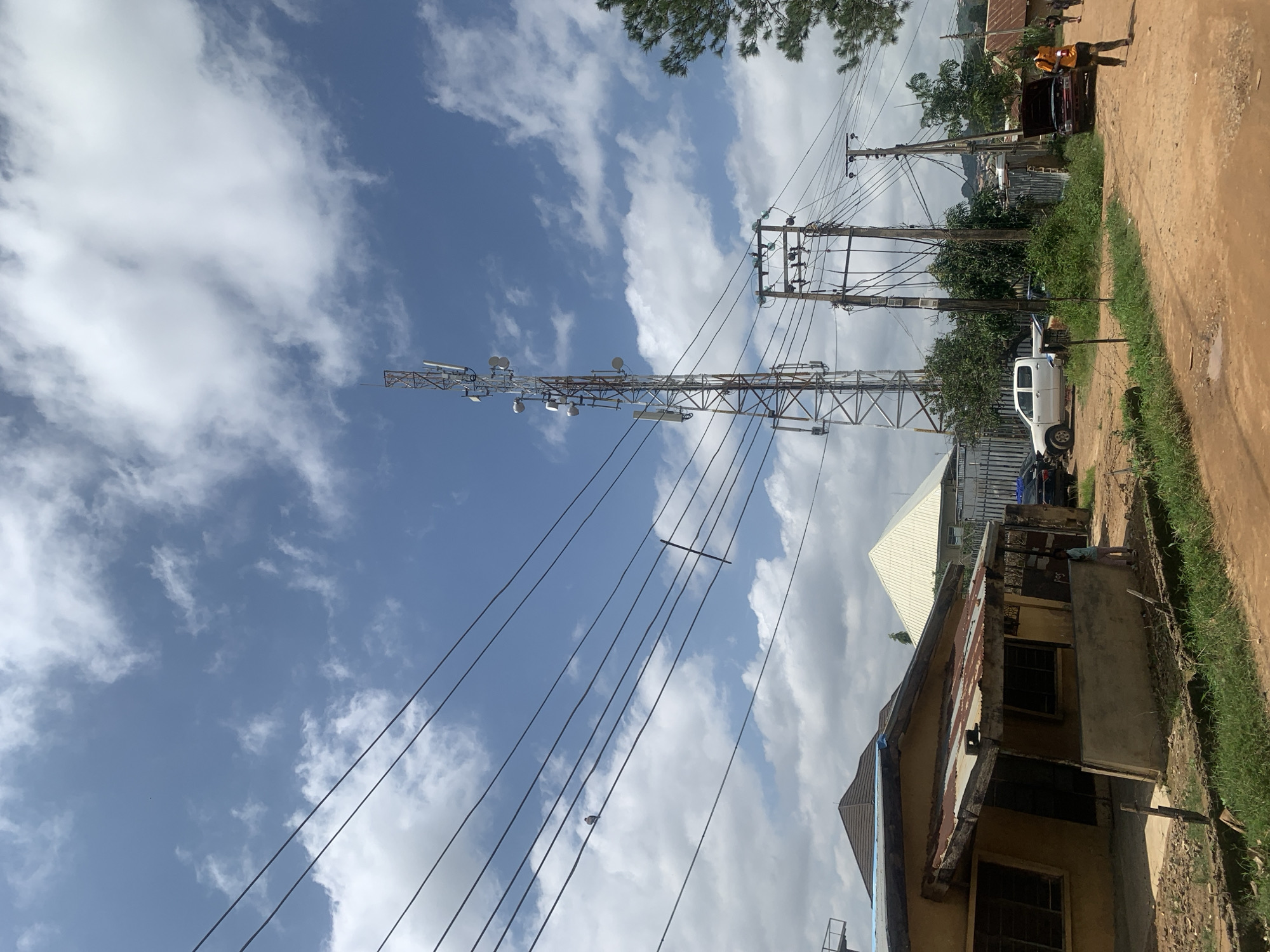 The image depicts a scene in Rafin Par, Nigeria, under a partly cloudy sky. A tall telecommunications tower with multiple antennas dominates the mid-ground, with power lines stretching across the frame. In the foreground, a dirt road runs past a weathered, yellow building with a rusted tin roof. To the right of the road, several vehicles are visible, including a white pickup truck, a car with its hood open, and other parked cars. A few people are present, including a figure in an orange shirt walking away from the camera on the right side of the road. The environment appears to be a semi-urban or rural area, with vegetation and other structures, including houses, visible in the background.
