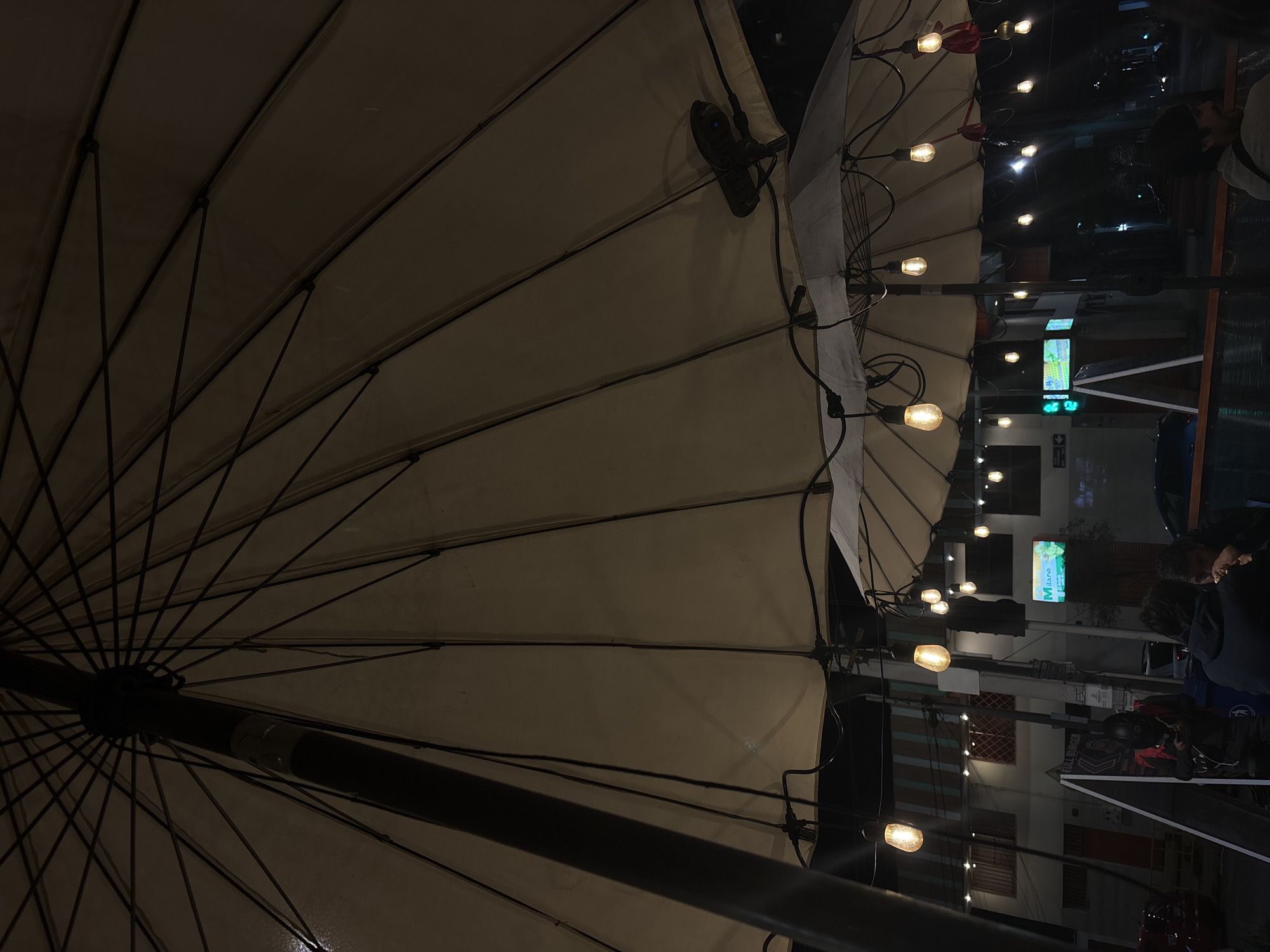 The image captures an outdoor seating area beneath large umbrellas, likely a restaurant or cafe, at night in Iquique, Chile. The umbrellas are illuminated by strings of Edison-style bulbs, casting a warm glow. In the background, a building with striped awnings and various windows is visible. Signs for "FULL BURGER" and "Milano" can be seen, indicating commercial establishments. Several people are present: one individual in a helmet and black and red jacket stands near a sign, looking at a phone; two individuals are seated at a table, one eating, and another person with a beanie is partially visible to the right. The scene appears to be on a street, with streetlights and building lights contributing to the nighttime ambiance.