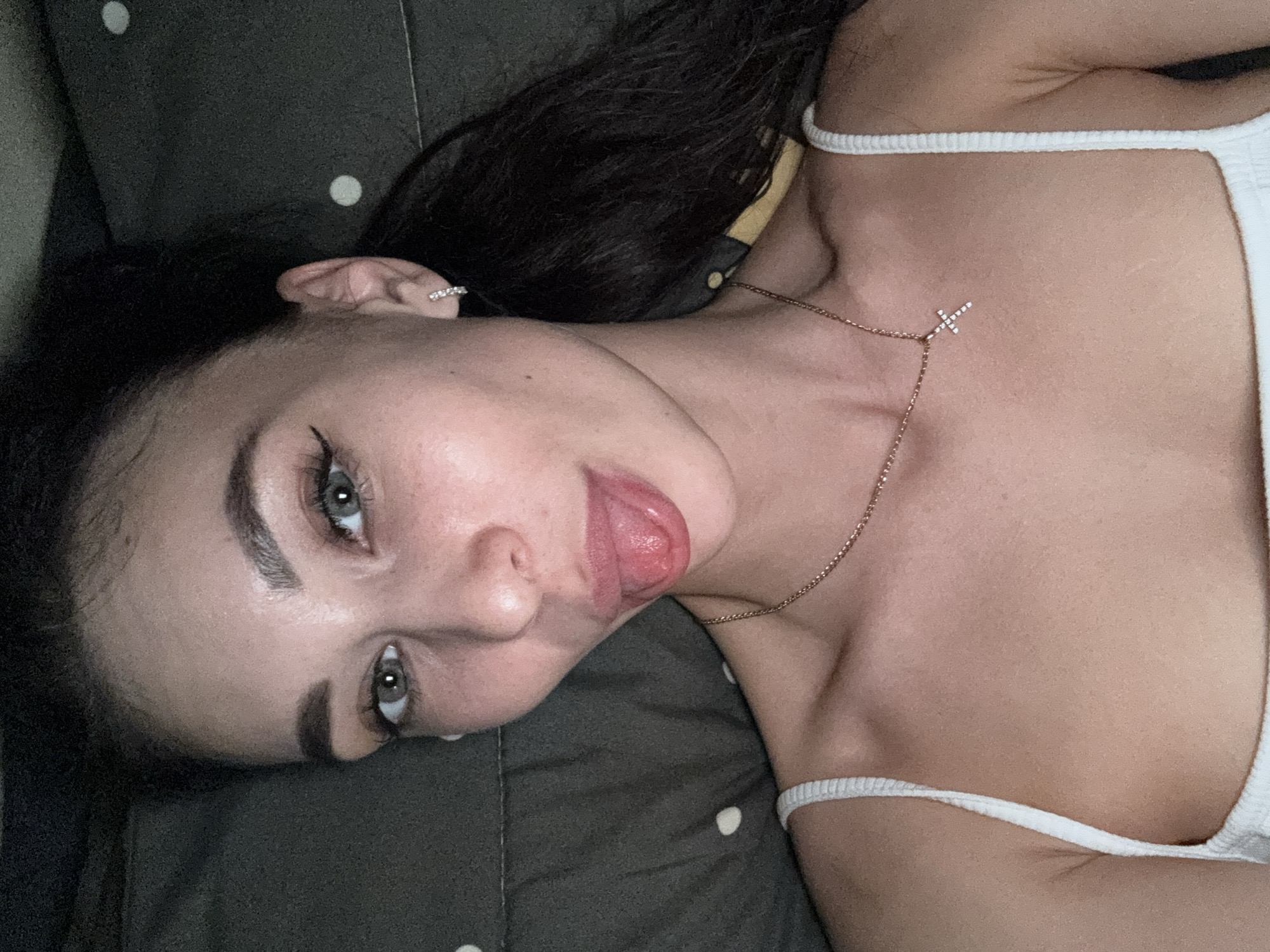 A fair-skinned young woman is lying on a dark gray surface with white polka dots. She is wearing a white tank top and a thin gold necklace with a small cross pendant. Her dark hair is pulled back. She has gray eyes, winged eyeliner, and is sticking out her tongue. An earring is visible in her left ear. The image was likely taken indoors. There are no indicators to prove the image was taken in the city of Chaikovsky, Russia.