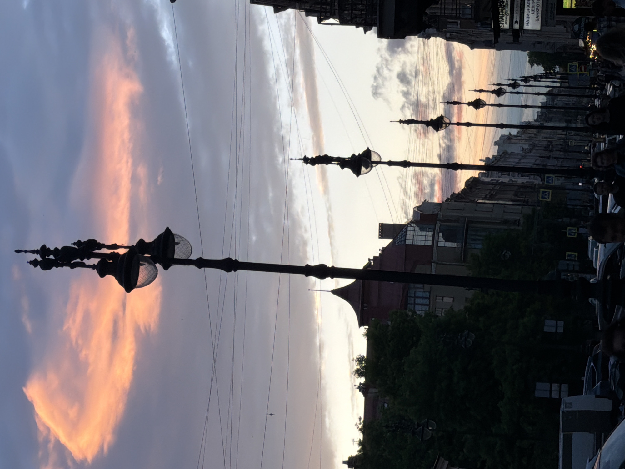 This is a street-level, eye-level, full shot of Nevsky Prospect in Saint Petersburg, Russia, captured at dusk. The sky is painted with hues of pink, orange, and purple as the sun sets, with scattered clouds adding texture. The foreground is dominated by a series of ornate black lampposts, each topped with multiple globe-like lights, lining the street.
On the left side of the image, a dense canopy of dark green trees partially obscures buildings with distinctive pointed red roofs and ornate architecture, typical of Saint Petersburg. Further down the street, on the right, more buildings with classical facades are visible, some featuring prominent signage, including what appears to be an advertisement for "НАЦИОНАЛЬНАЯ СЛУЖБА БАНКРОТСТВА" (National Bankruptcy Service).
The street itself is busy with traffic, including cars and a van. Several pedestrians are visible, their forms silhouetted against the twilight. Some are walking along the sidewalk, while others appear to be gathered near the street. The overall impression is a vibrant urban scene transitioning from day to night, with the iconic architecture of Saint Petersburg providing a dramatic backdrop to the bustling activity.