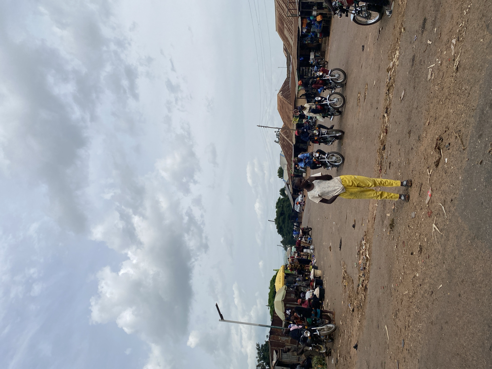 In Turu, Nigeria, on a dusty, unpaved road, a young boy stands in the foreground wearing a white t-shirt and bright yellow trousers. The scene depicts a bustling marketplace under a cloudy sky. Several motorcycles, some with riders, are present on the road. In the background, a row of market stalls with corrugated metal roofs line the street. People are gathered around the stalls, some appearing to be vendors and others customers. The environment is characterized by the presence of dirt, debris, and scattered items on the ground, typical of a lively, open-air market. The overall atmosphere suggests a busy day at the market.