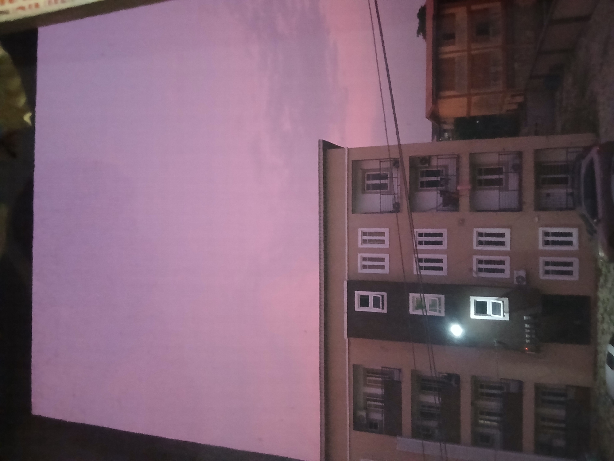 A photograph captures a dusky sky with hues of pink and purple, suggesting either dawn or dusk. In the foreground, a multi-story apartment building with beige and dark gray facade dominates the scene. The building features numerous windows, some illuminated by interior lights, and balconies with black railings. On the right side of the image, a smaller, orange-roofed building is visible. Power lines crisscross the frame. The lower portion of the image shows the top of a dark-colored car parked in front of the apartment building. The setting appears to be an urban environment, possibly in the city of Unknown, Unknown.