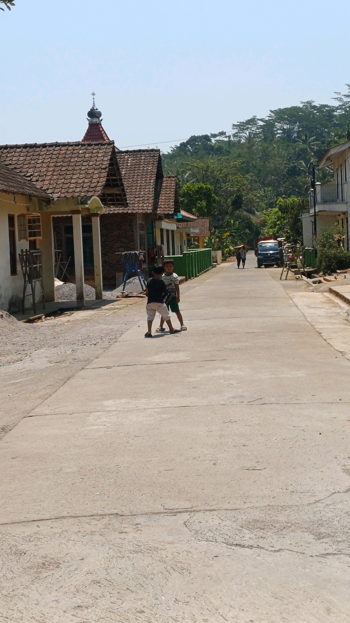 In Pringsurat, Indonesia, two young boys stand on a paved street. One boy is wearing a black t-shirt and white shorts, while the other wears a striped t-shirt and green shorts. They appear to be facing each other, possibly interacting. The street is lined with houses on either side, some with tiled roofs and unfinished sections. A small mosque with a dome and spire is visible on the left. In the background, trees and vegetation cover a hillside. A dark-colored vehicle is parked further down the street, and two adults are walking in the distance. The scene is set under a clear, bright sky.