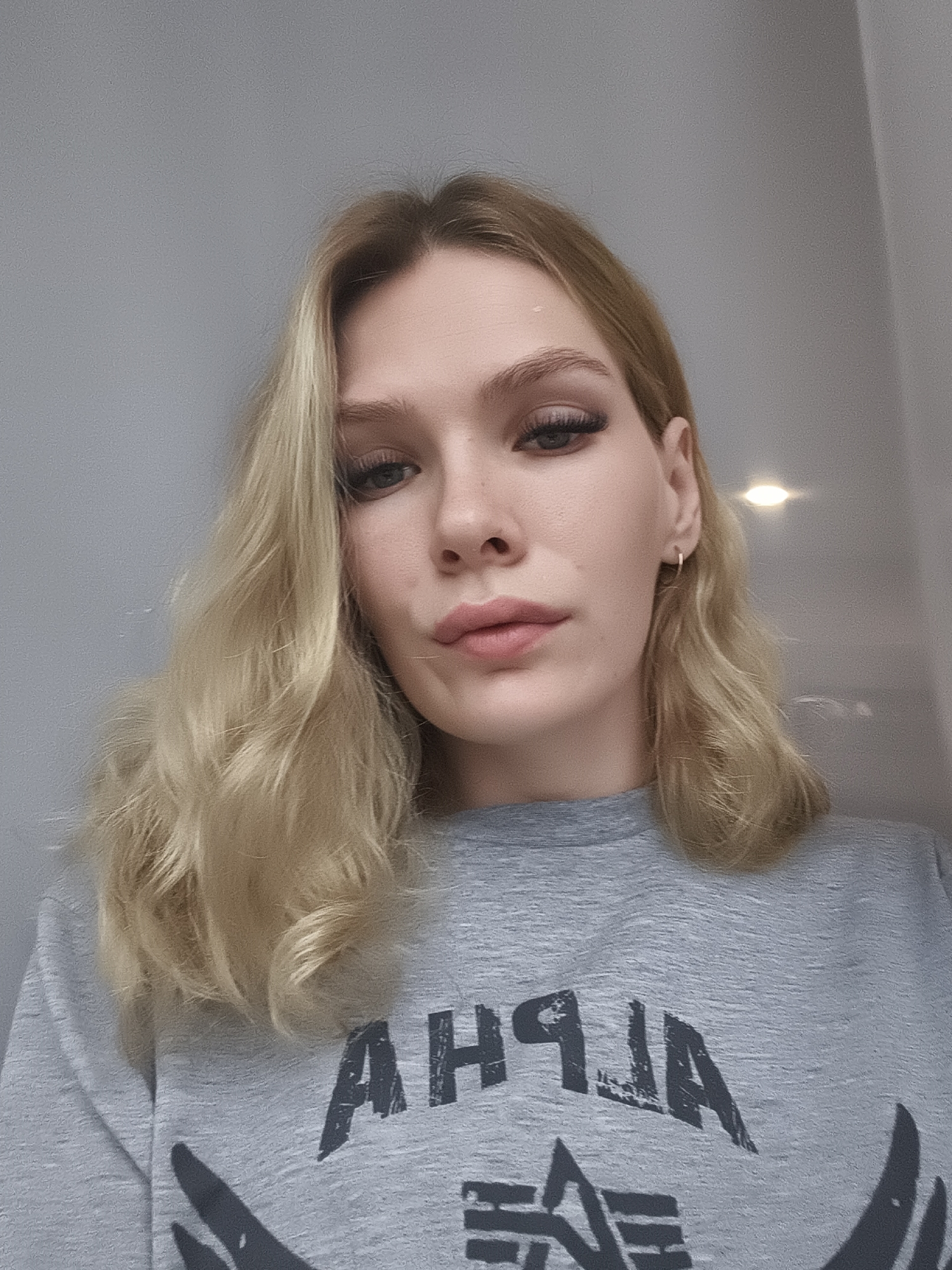 A head-and-shoulders shot of a woman with blonde wavy hair wearing a gray long-sleeved shirt with the word "ALPHA" printed on it in a stylized font. She has fair skin, defined eyebrows, and full lips. She is facing the camera, with a neutral expression. The background is a plain gray wall.
The location Русский Вожой in Russia is not identifiable in the image.