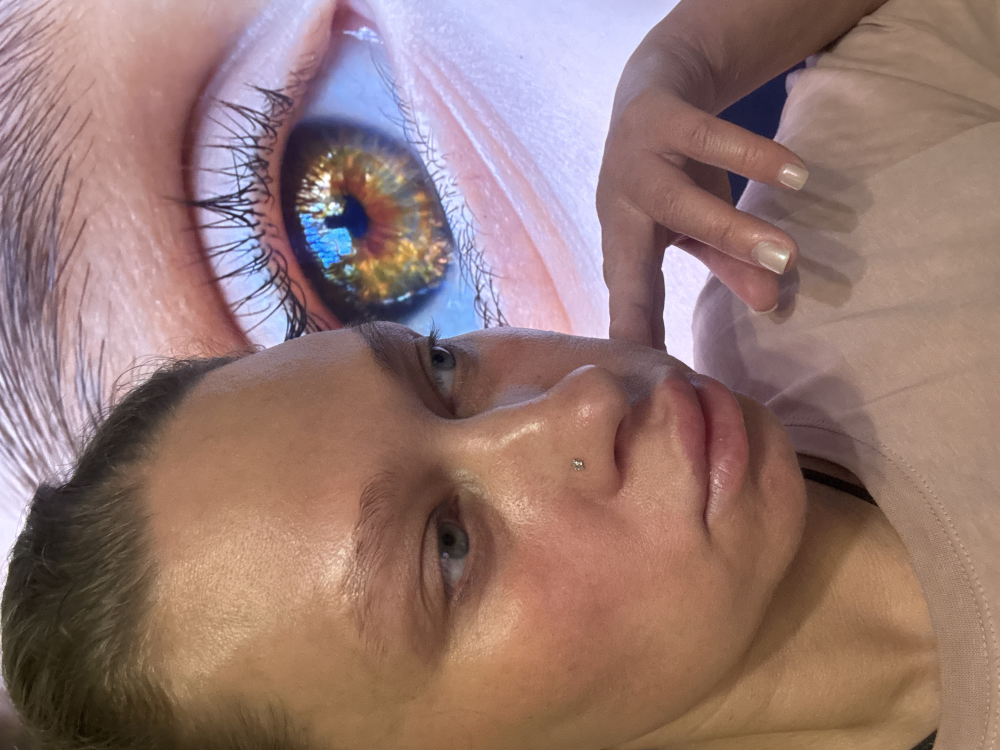 A close-up shot of a woman's face with a nose piercing is juxtaposed with a large image of a human eye in the background. The woman is positioned at an angle, her gaze directed upwards. Her hand is visible in the upper right, positioned as if touching the background eye. The background features a giant eye in focus with an iris that reflects a cityscape, though it's difficult to discern specific architectural details, it is not identifiable as Perm, Russia. The foreground shows the woman's face in detail, revealing skin texture and slight imperfections. She is wearing a light pink shirt.