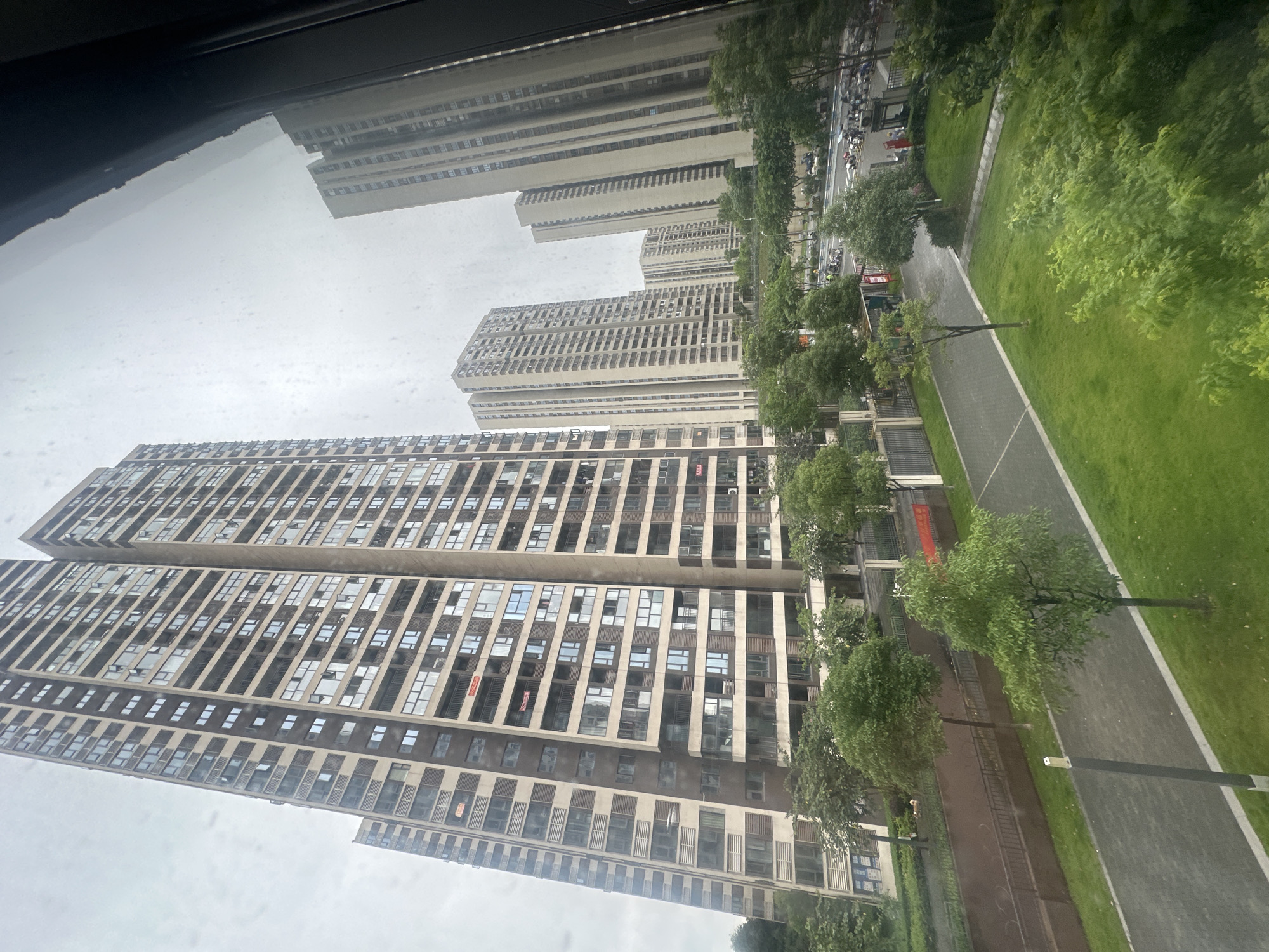 This image depicts a view of a residential area in the Chongqing High-tech Zone, China, on a cloudy day. The foreground features a neatly manicured lawn with a paved walkway and a few young trees. Beyond the lawn, there is a road with several motorcycles parked on the side and some passing traffic. A red banner is visible on a fence. In the background, towering residential buildings dominate the skyline. These are modern, multi-story apartment complexes with numerous windows. The sky is overcast, casting a diffused light over the scene. The overall impression is of a densely populated urban environment with green spaces interspersed among the high-rise buildings.