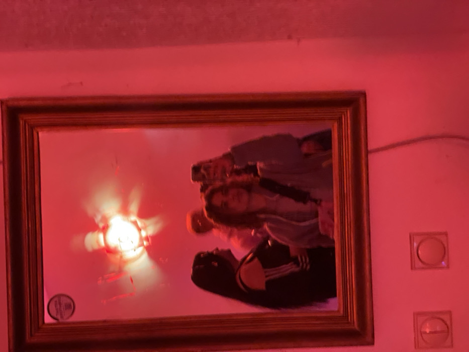 A framed mirror reflects a group of people, including a person wearing Adidas-striped clothing, along with a bright light fixture. The room is bathed in red light. The frame has a golden hue. A light switch is partially visible on the wall to the right of the mirror.