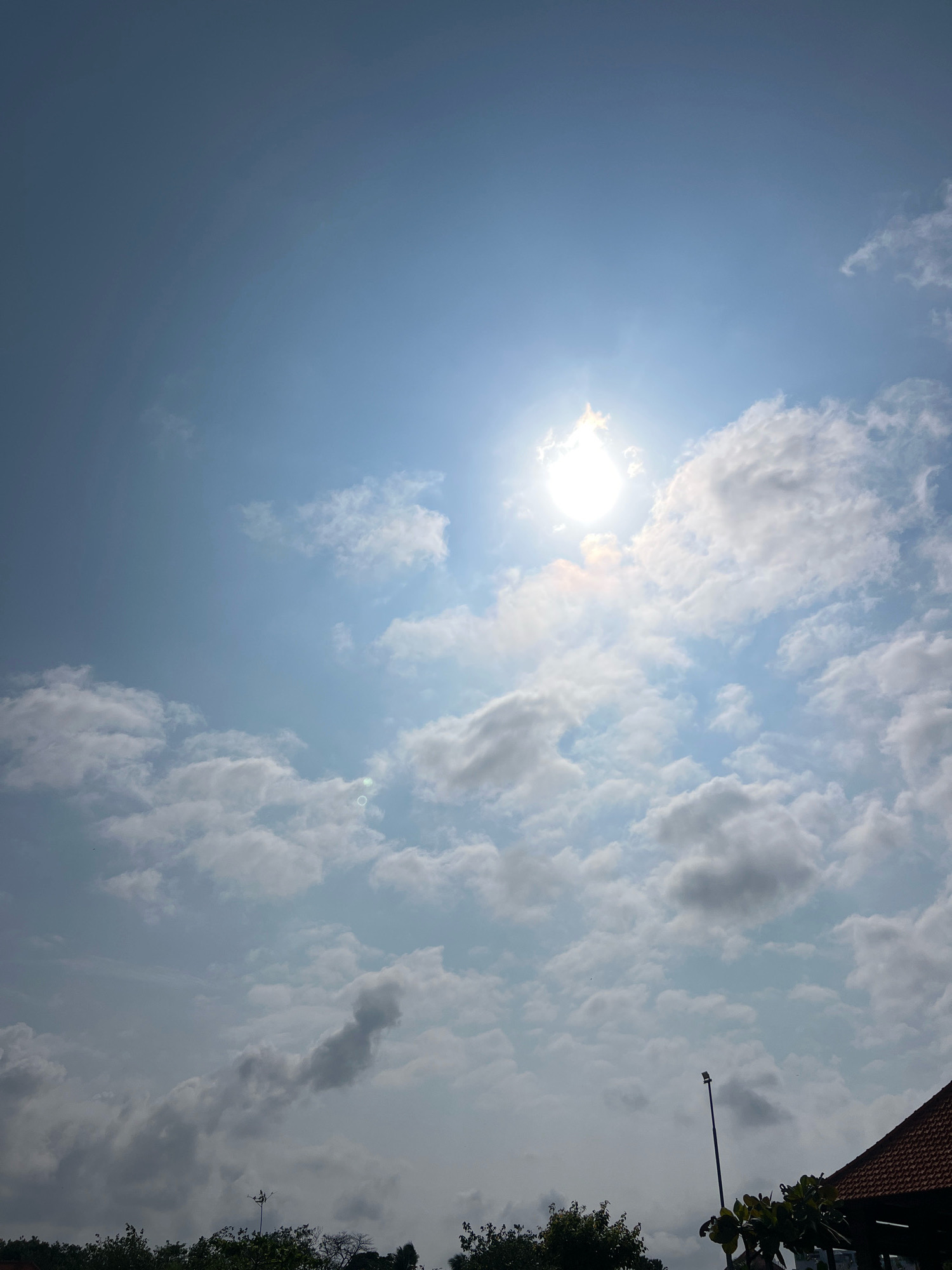 The image captures a bright, partly cloudy sky over Kuta, Indonesia. The sun is high in the sky, casting a strong glare and creating lens flares. Fluffy white clouds are scattered across the light blue expanse. In the foreground, the tops of lush green trees are visible, hinting at a tropical environment. A portion of a building with a terracotta tiled roof is present on the right side of the frame, along with a dark lamppost rising from behind the foliage. The scene appears to be an outdoor view, possibly from a rooftop or elevated position, looking towards the sky on a sunny day.
