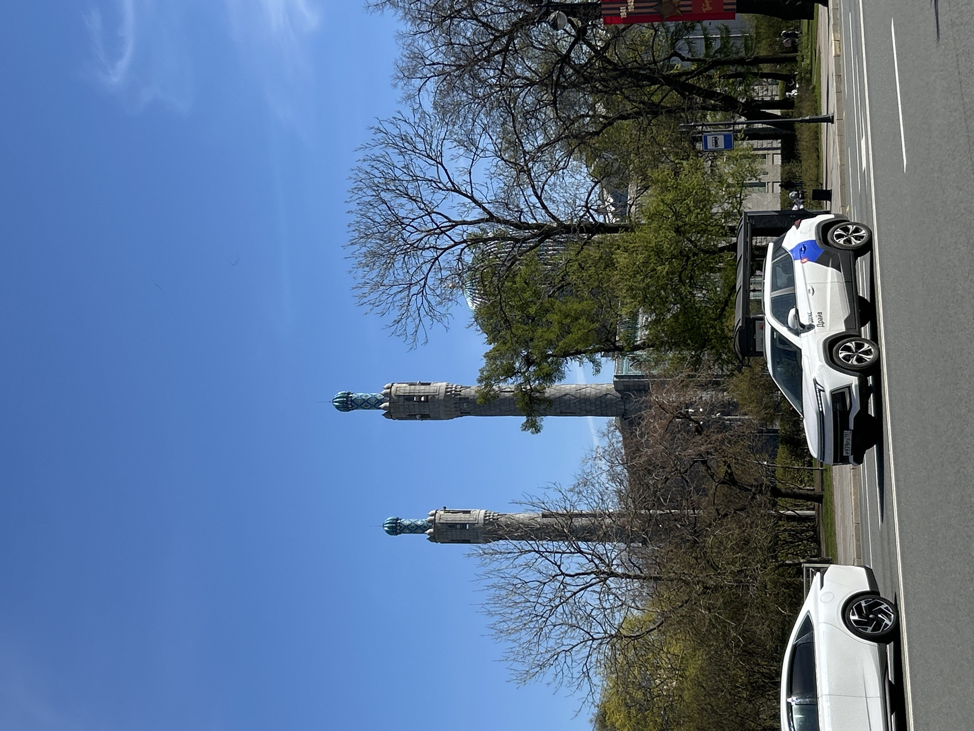 The image depicts a street scene in Saint Petersburg, Russia, featuring the Rostral Columns as viewed from the road. Two white cars are parked along the side of the road, occupying the right side of the frame. The Rostral Columns, positioned in the center of the image, rise against a clear blue sky. Bare trees and green foliage are visible around the base of the columns. In the background, a banner is visible along with a bus stop. People appear to be walking in the background behind the bus stop.