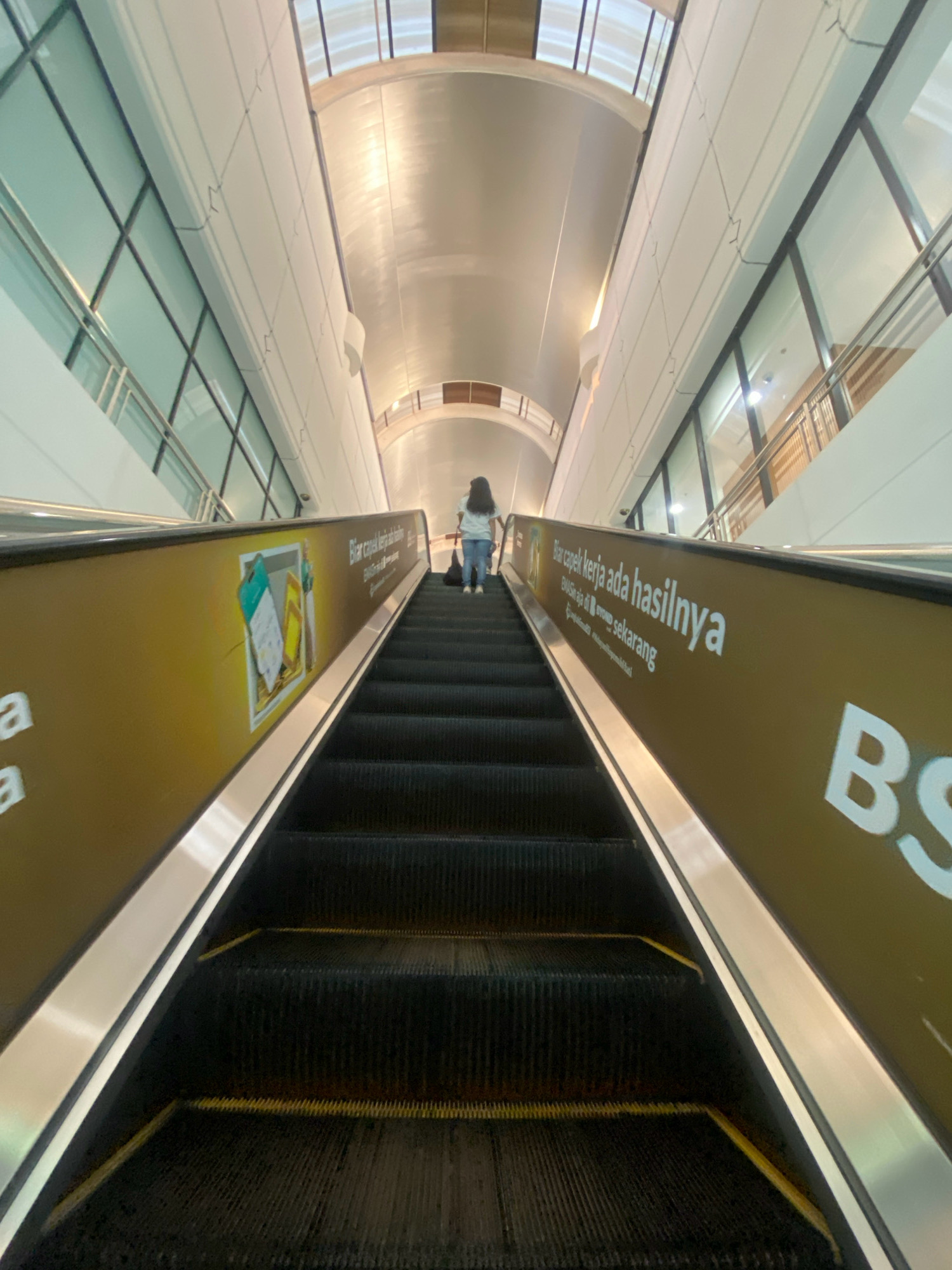 A low-angle, upward perspective shot shows a person ascending an escalator within a modern, brightly lit indoor environment, possibly an airport terminal or a large shopping mall in Batam, Indonesia. The escalator is black with metallic handrails and yellow safety strips at the edges of each step. A large, gold-colored advertisement wraps around the side of the escalator, featuring text in Indonesian, including "Biar capek kerja ada hasilnya" (Let tired work have results) and "Emasin aja di ovo sekarang" (Gold it on ovo now). The architecture of the building is visible in the background, with a high, curved ceiling and large glass windows on the sides, revealing an external structure. The person on the escalator is wearing a light-colored t-shirt and blue jeans and is seen from behind, carrying a dark bag.