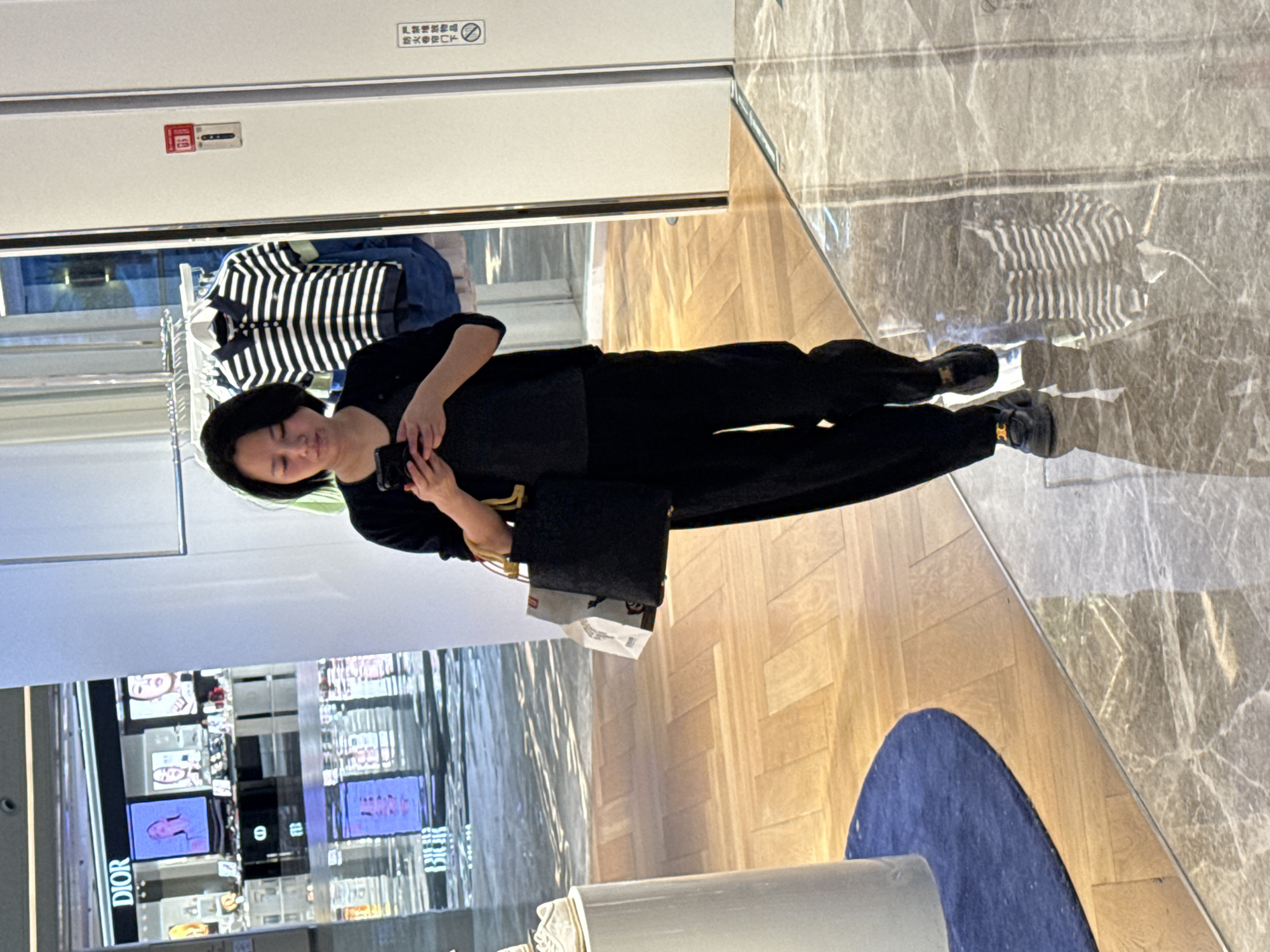 A woman stands indoors, possibly in a retail environment, holding a mobile phone. She is dressed in black clothing and is holding a black bag. The background includes a display with "DIOR" branding, and a clothing rack with apparel. The location appears to be a store in a shopping mall in the city of Liangjiang, China.