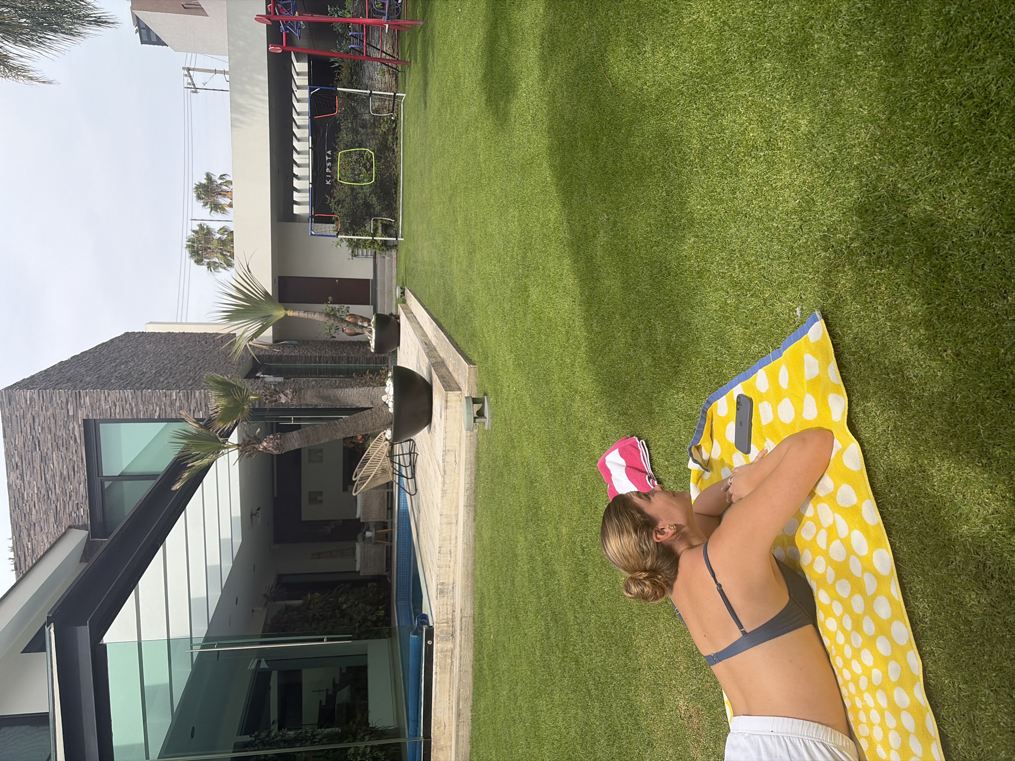 A woman is sunbathing on a yellow and white polka-dot towel on a green lawn. She is wearing a grey swimsuit top and white shorts. A pink and white striped towel is near her head. In the background is a modern house with a black roof and large glass windows, surrounded by palm trees. A swimming pool runs along the side of the house. Further back, there's a children's play structure and a soccer net.