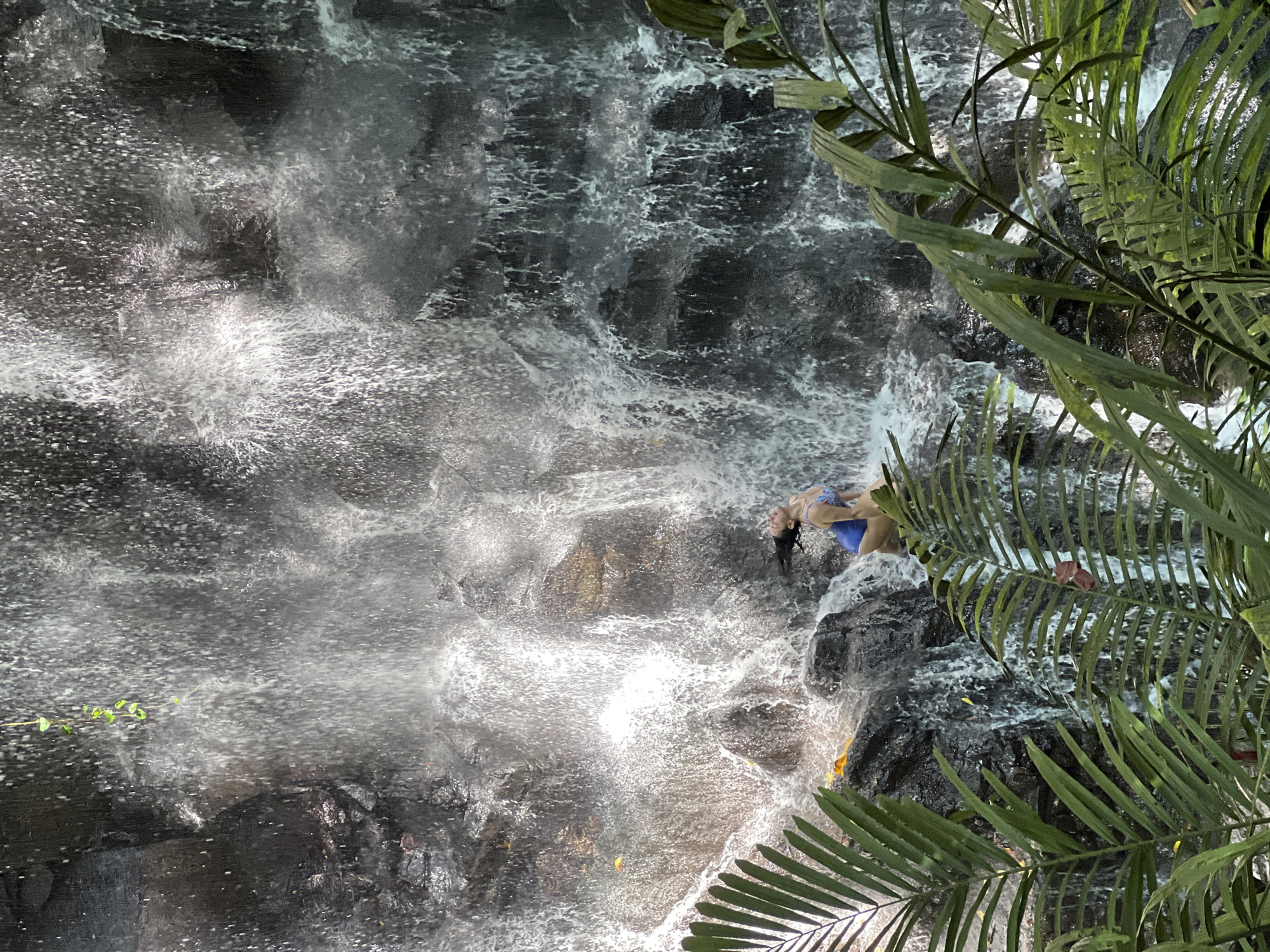 The image captures a powerful waterfall cascading over dark, rugged rocks in a lush, tropical environment, consistent with the location context of Gianyar, Indonesia. The water is highly aerated and white with froth and spray as it tumbles downwards.
On a rocky ledge to the right of the main waterfall, two people are visible. One individual, wearing a blue bikini or swimsuit, is lying on their back with their head tilted upwards, appearing to be enjoying the mist and the force of the water. Their dark hair is wet. Partially obscured behind this person, another individual in dark clothing, possibly a wetsuit or dark swimwear, is visible, standing or kneeling closer to the cascading water.
The activity suggests a recreational or adventure experience at the waterfall, such as enjoying the natural environment, bathing, or participating in a water-based activity. The scene is brightly lit, indicating it is daytime. Green, palm-like foliage frames the right side of the image, emphasizing the natural, jungle-like setting. No visible text is present in the image.