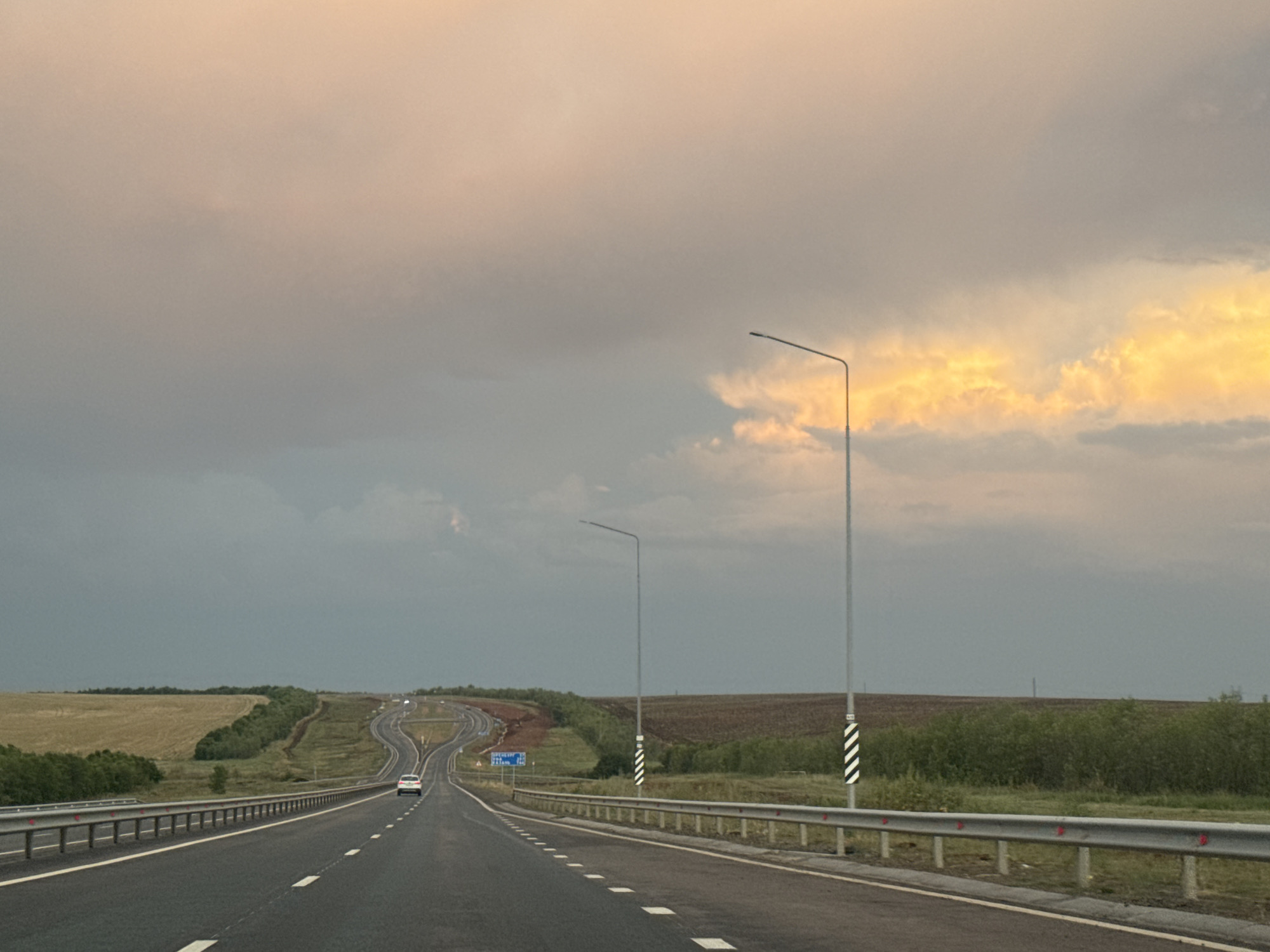 The image depicts a highway with a car driving on it, likely in or near the city of Syrt, Russia. The sky is overcast with some areas of bright, yellowish clouds, suggesting either sunrise or sunset. The road is asphalt, with white lane markings and a metal guardrail on the right side. Lampposts are visible along the road. The surrounding landscape consists of fields, trees, and rolling hills. The overall impression is of a rural highway scene during twilight.