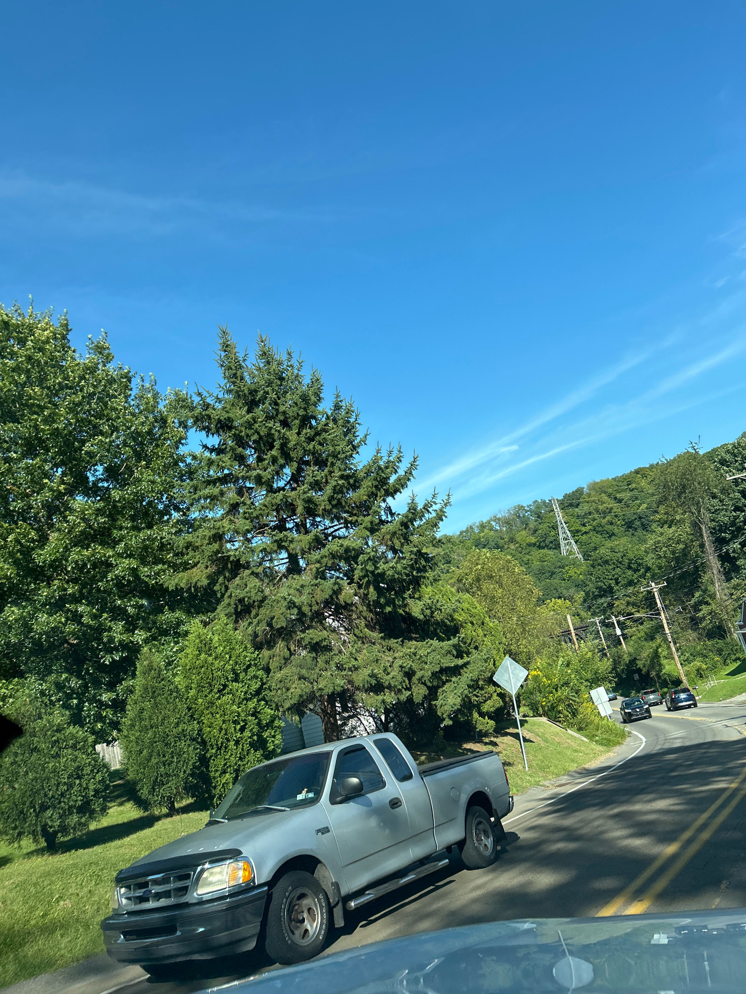 The image depicts a bright, sunny day in Collier Township, United States of America, with a clear blue sky and some wispy clouds. A silver pickup truck is parked on the grassy shoulder of a paved road, which curves to the right. The truck has a black bug deflector on the hood and appears to be a Ford F-150. There are trees and lush green foliage on either side of the road, with a hill covered in trees in the background. A tall, lattice-style transmission tower is visible on the hill. Further down the road, two dark-colored cars are driving away from the camera. A diamond-shaped road sign is posted on the left side of the road, near the pickup truck. The scene suggests a rural or suburban setting with natural beauty.
