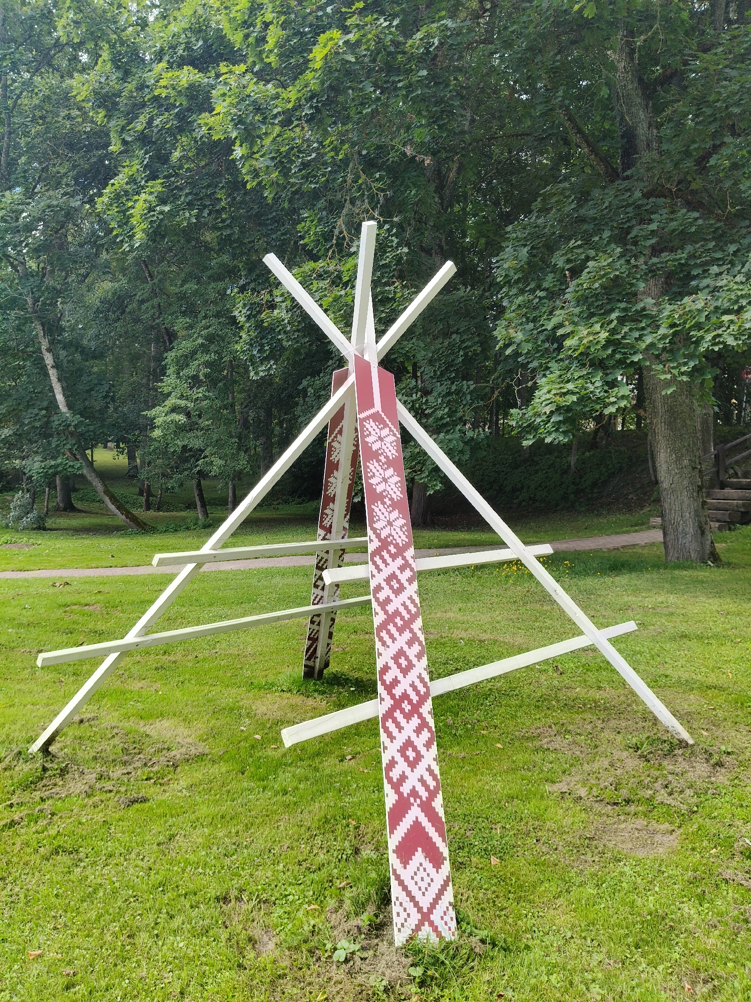 In Koknese, Latvia, a structure with a tripod-like base made of white wooden beams stands on a grassy area. A red and white patterned banner is attached to the central part of the structure. The background is dominated by lush green trees, suggesting a park or forest setting. The ground is covered in grass, with some areas of exposed soil. The overall scene depicts an outdoor installation within a natural environment.