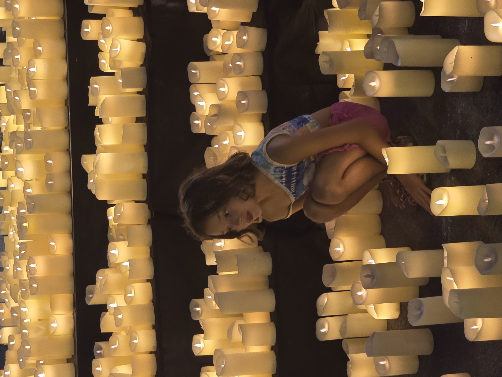 A young girl is crouching amongst a large display of flickering, battery-operated candles. The candles are arranged on multiple levels and on the floor, creating a warm, ambient glow. The girl, wearing a tie-dye top and pink shorts, looks down at the candles with an inquisitive expression. The scene appears to be an indoor event or installation, possibly for a memorial or a festive occasion, with a dark, neutral background that emphasizes the light from the candles. The location is identified as Sant Adrià de Besòs, Spain.