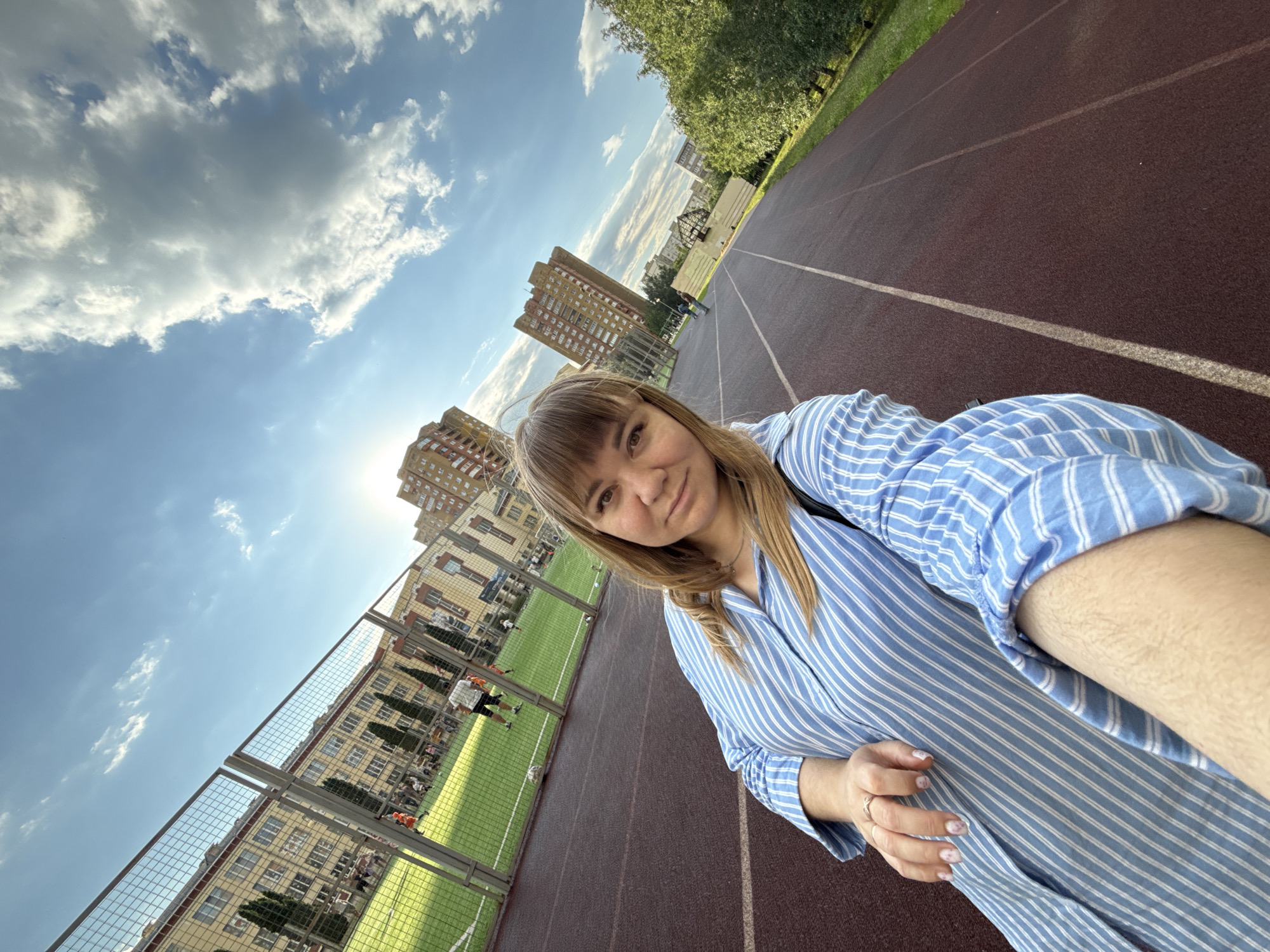 A woman with blonde hair and bangs takes a selfie while standing on a running track. She is wearing a blue and white striped shirt. In the background, there is a green sports field enclosed by a metal fence, a building with many windows, trees, and a cloudy blue sky. The sun is visible in the sky, suggesting it is daytime. The image appears to have been taken outdoors in an urban environment, possibly a school or public park. The woman has a neutral expression. The running track has red-brown surfacing and white lines.