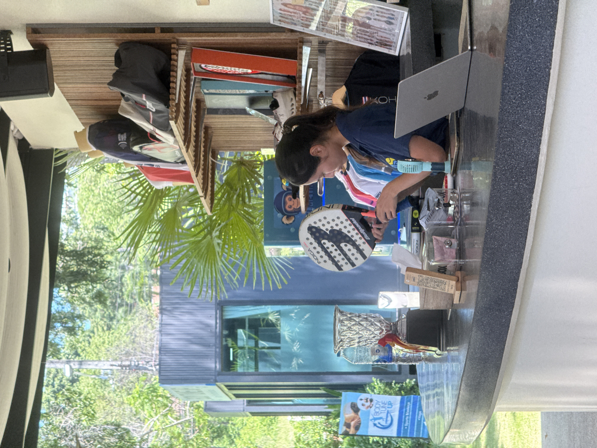 A woman with dark hair pulled back in a ponytail, wearing a navy blue shirt, stands behind a curved, dark grey counter. She is holding a white and black padel racket, examining it. In front of her on the counter is a silver laptop, a small blue fan, a mobile phone, and various other items including a trophy, bottles of water, a QR code stand, and tissues. Behind her, a wooden shelf unit displays several bags and apparel items. The scene is outdoors, with lush green trees and foliage visible in the background, suggesting a tropical or subtropical environment. A blue banner with the text "BODY TUNE UP" is partially visible to the left of the counter. The overall setting appears to be an outdoor sports facility or a recreational area.