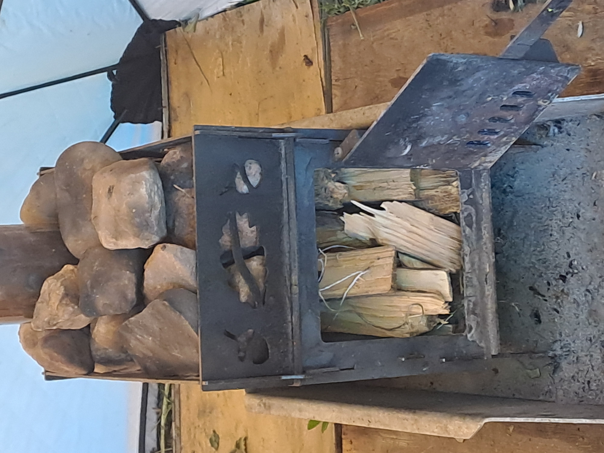 This image captures a rustic outdoor or semi-outdoor scene, centered around a compact metal wood-burning stove. The stove, dark gray or black, has an open hinged door on its top, revealing split logs neatly stacked inside, ready for burning. The stove features decorative cutouts on its visible side, possibly depicting a leaf or organic shapes.
To the left of the stove, a pile of sizable, rounded stones is stacked on a wooden platform, possibly serving as a heat reservoir or part of a sauna heating system. The ground or floor around the stove is composed of wooden planks and some loose dirt or ash.
In the background, a light blue fabric structure, likely a tent or tarp, is visible, with a dark, folded item (possibly fabric or a bag) hanging from a bar. The overall setting suggests a temporary dwelling or a campsite, possibly in a wilderness area. The lighting indicates it is daytime. There are no people visible, and no discernible text. The scene conveys an atmosphere of preparedness for heating or cooking in a simple, outdoor environment. The location context provided is Полянки, Russia.