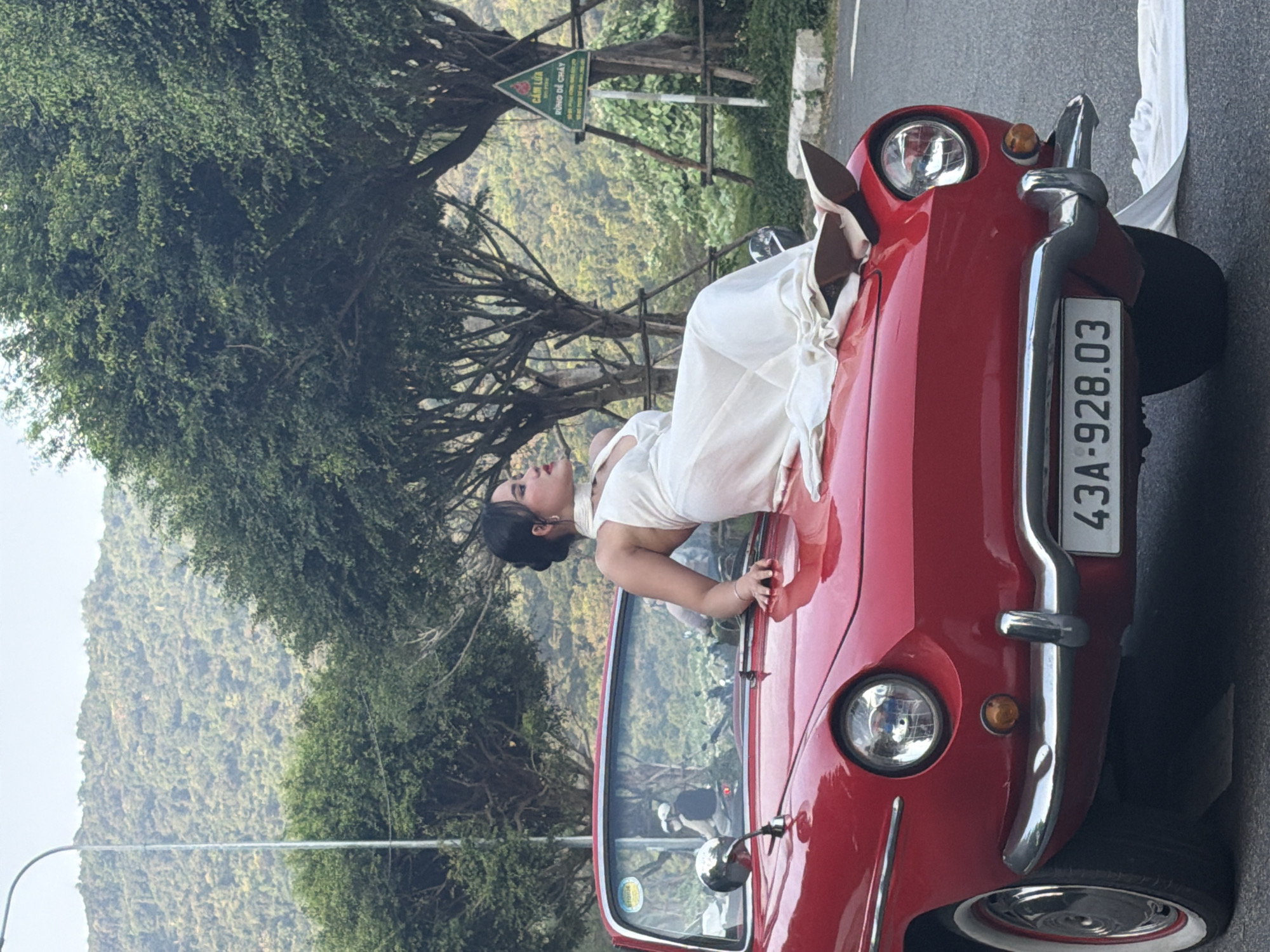 A woman in a white dress leans out of the driver's side window of a vintage red car. The car has a license plate that reads "43A-928.03". The setting appears to be a mountainous area with dense foliage and trees. A street lamp is visible on the left side of the image. A road sign is seen in the background. The location is in Đà Nẵng, Vietnam.