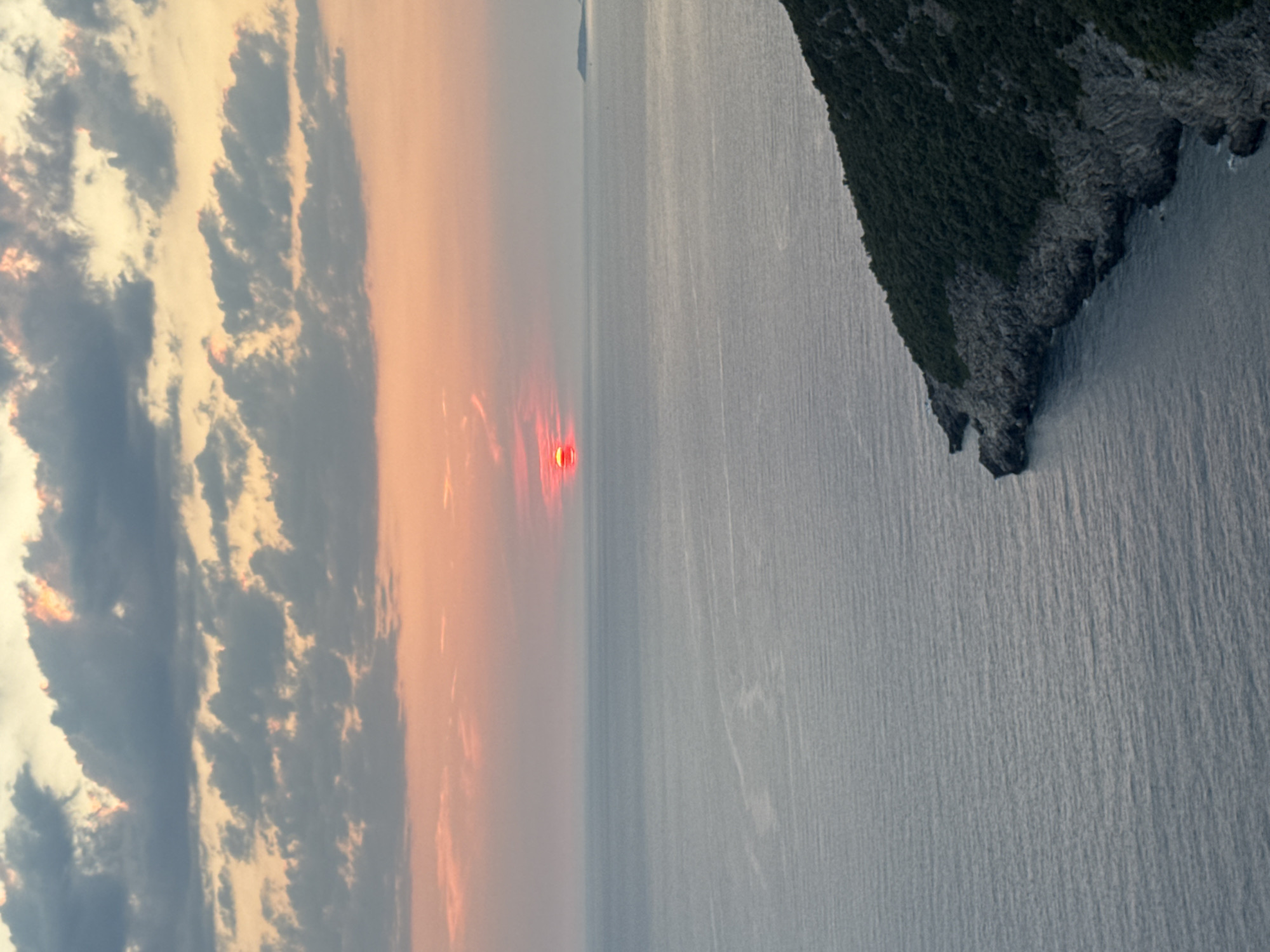 The image depicts a serene coastal sunset in Zagora, Montenegro. The sky is a gradient of warm hues, transitioning from pale pink and orange near the horizon to soft blues and grays higher up, punctuated by scattered clouds. The sun, a vibrant red-orange orb, is partially obscured by a thin layer of clouds just above the horizon, casting a faint glow on the calm sea. The water itself is a deep, muted blue, reflecting the sky's colors with subtle ripples. In the foreground, a rocky, tree-covered peninsula juts out into the sea, its dark green foliage contrasting with the lighter tones of the sky and water. The rugged coastline is characterized by steep cliffs and rocky formations. The scene is devoid of people or any discernible human activity, emphasizing the natural beauty of the location. The overall impression is one of peaceful tranquility as day transitions into evening.