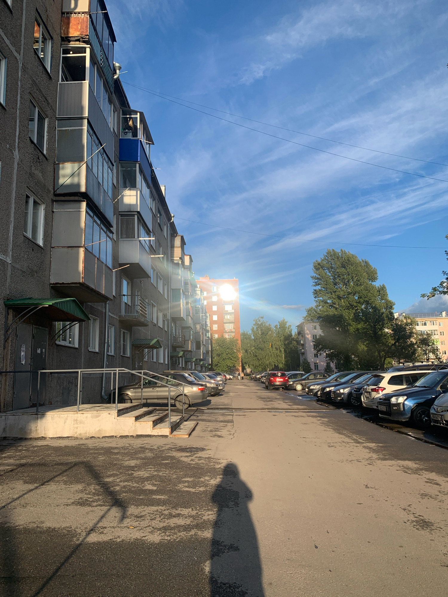 The image depicts a residential courtyard in Prokopyevsk, Russia. A multi-story Soviet-era apartment building with balconies lines the left side of the frame. The building's facade is rough and gray, with some balconies showing signs of age and modification. To the right, a row of parked cars stretches down an asphalt lot. The sun is low in the sky, casting long shadows and creating a lens flare effect. The sky is blue with wispy clouds. A large, mature tree stands in the middle ground, and other buildings are visible in the background. The ground is wet in places, suggesting recent rain. A person's shadow is prominently cast on the ground in the foreground.