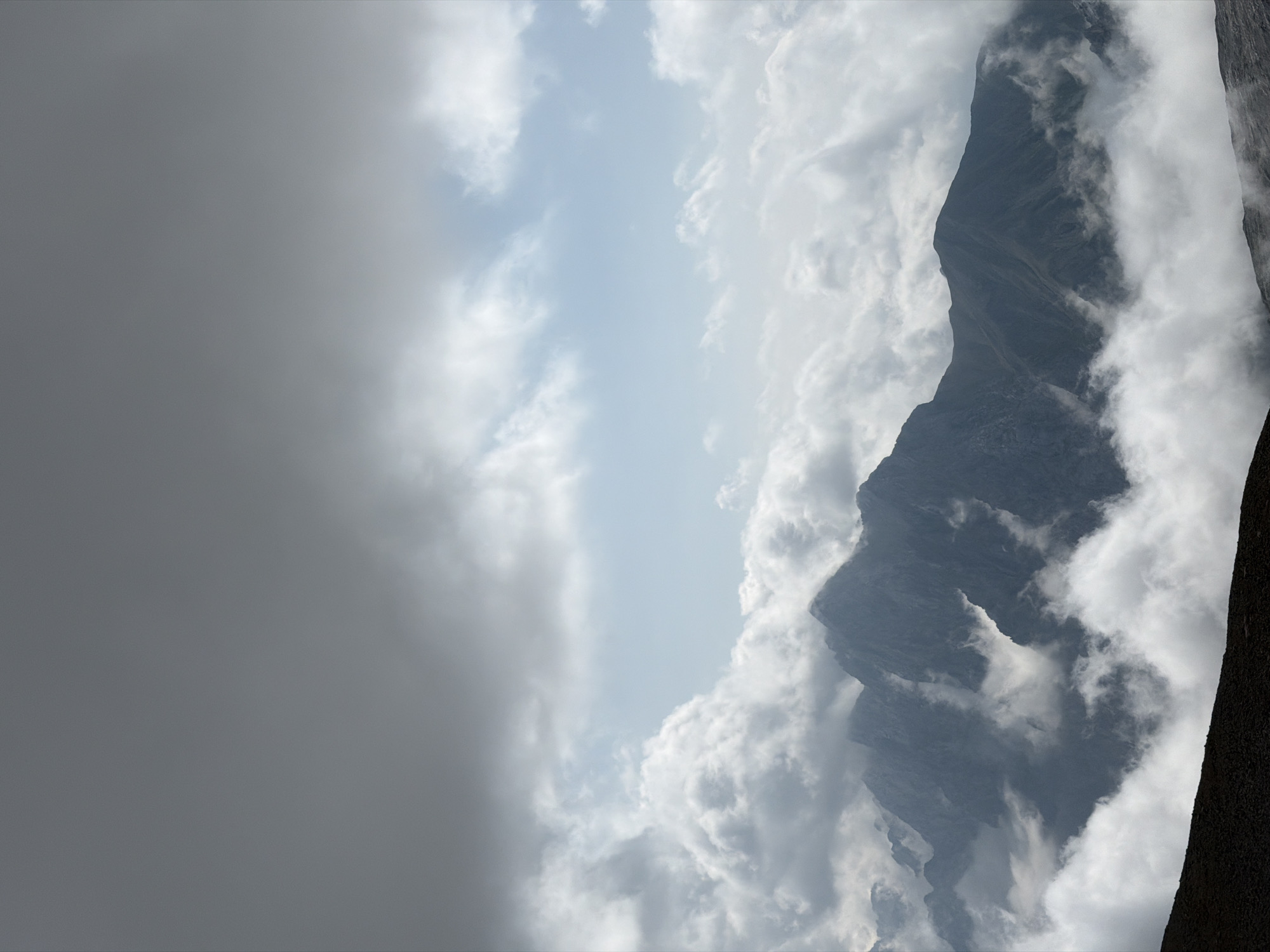 The image depicts a majestic mountain range shrouded in dramatic clouds. Dark, heavy clouds dominate the upper portion of the frame, with patches of lighter, scattered clouds interspersed with a pale blue sky in the middle ground. Below these, a dense layer of white, fluffy clouds hugs the peaks of the mountains, obscuring their bases and creating a sense of ethereal grandeur. The mountains themselves are rugged and dark, with sharp, angular forms indicative of a rocky, alpine environment. The lower foreground shows a dark, barren slope, possibly scree or exposed earth, providing a stark contrast to the misty heights. The scene is one of natural, untamed beauty, evoking a feeling of awe and isolation. The image does not contain any people, man-made structures, or specific identifiable landmarks that would place it within Garmisch-Partenkirchen, Germany, or any other specific urban location.