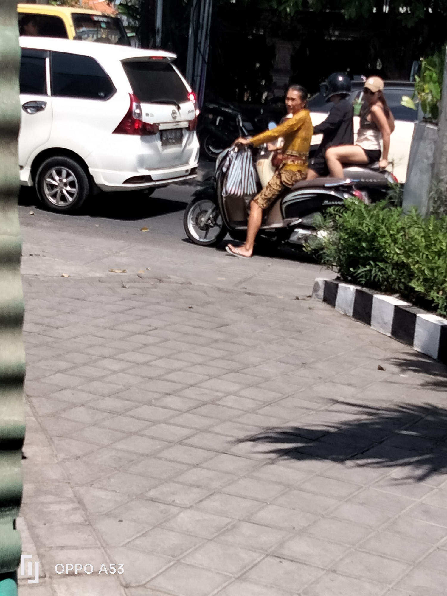 In North Kuta, Indonesia, on a paved road with a tiled surface, a man wearing traditional Balinese clothing, a yellow long-sleeved shirt and patterned pants, rides a scooter. He is wearing flip-flops and has a bag with striped fabric hanging from the handlebars. Behind him, a woman with a tan hat is seated on the scooter. In the background, a white SUV is parked. The scene is lit by sunlight, casting shadows on the ground.