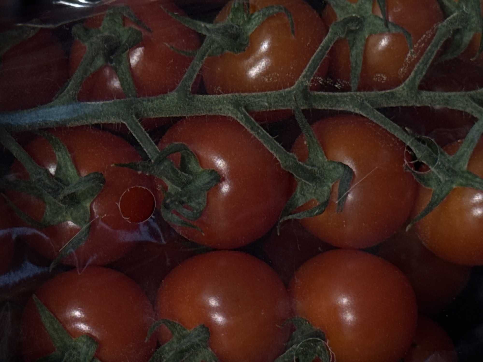 A cluster of ripe, red cherry tomatoes is visible, enclosed in clear plastic packaging. The tomatoes are attached to green stems, with some small green leaves also present. The background is dark and indistinct, suggesting an indoor setting or the interior of a package. There is no indication of the location being in Moscow, Russia, or any specific city. No people, actions, or interactions are depicted.