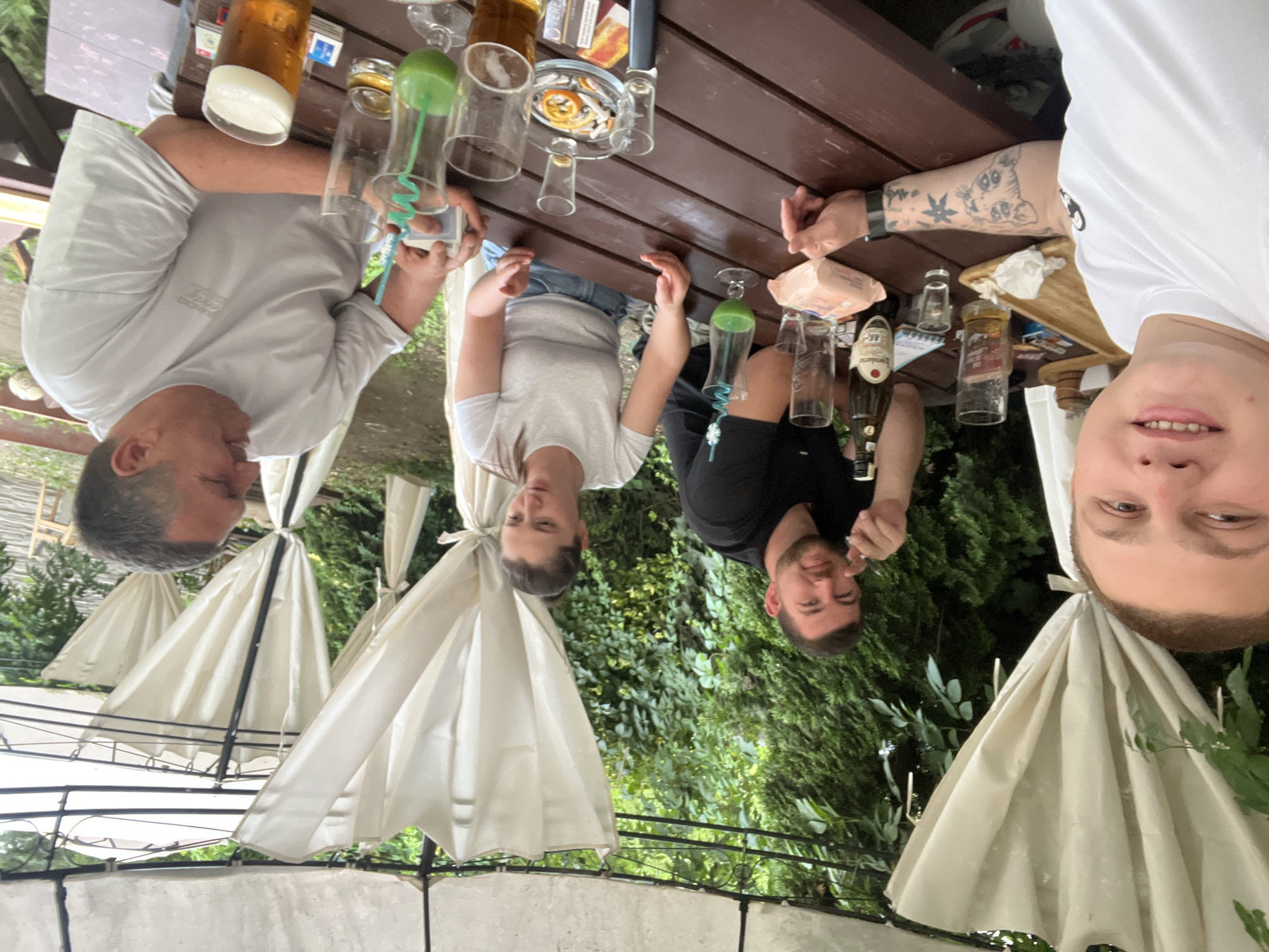 A group of four people, three men and one woman, are seated around a wooden table outdoors, beneath a white canopy. The man on the far left is taking a selfie, with his arm extended and his face close to the camera. He is wearing a white t-shirt and has tattoos on his arm. To his right, a man in a black t-shirt is looking towards the camera, holding a small object in his hand. Next to him, a woman in a white t-shirt and denim shorts is looking down at the table, with a green drink in front of her. The man on the far right, wearing a grey t-shirt, is looking to his left and smiling while holding a small white box. On the table are various drinks, including glasses of beer and green-colored beverages, a bottle of alcohol, shot glasses, a pack of wet wipes, an ashtray with cigarette butts, and some papers. The environment appears to be a beer garden or an outdoor dining area with lush green foliage in the background. There are white curtains hanging from the canopy.