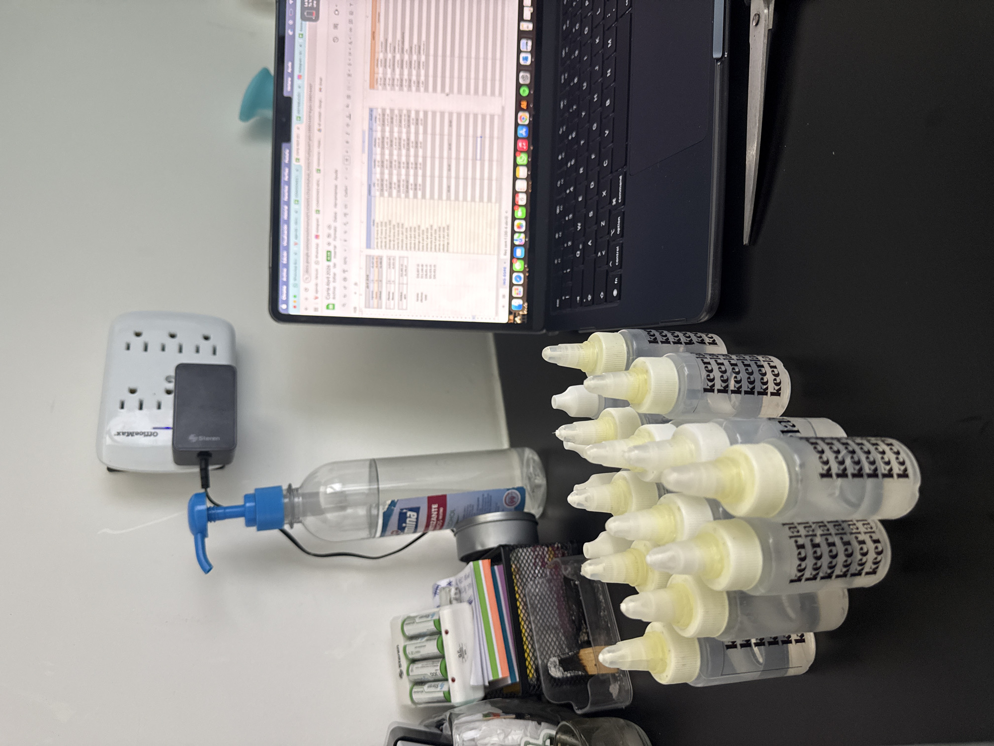 A top-down view reveals a cluttered desk surface bisected into two distinct areas: a clean, white section on the left and a darker, potentially black, section on the right.
On the left, an OfficeMax surge protector is plugged into the wall, and a black Steren adapter is connected to a clear spray bottle with a blue nozzle. The bottle is labeled "TIZANTE" and "mina."
Moving toward the center of the desk, there's a small metal organizer containing several AA batteries, colored note cards, and other office supplies.
On the right side of the desk and partially obscured by the organizer, there's a collection of small, clear bottles with white caps, labeled "keerlal." A laptop, partially open, displays a spreadsheet. The screen is on, and the keyboard is visible. A pair of metal tweezers or pliers lies next to the laptop.
There is no information about San Bernardino Tlaxcalancingo or people in the image.