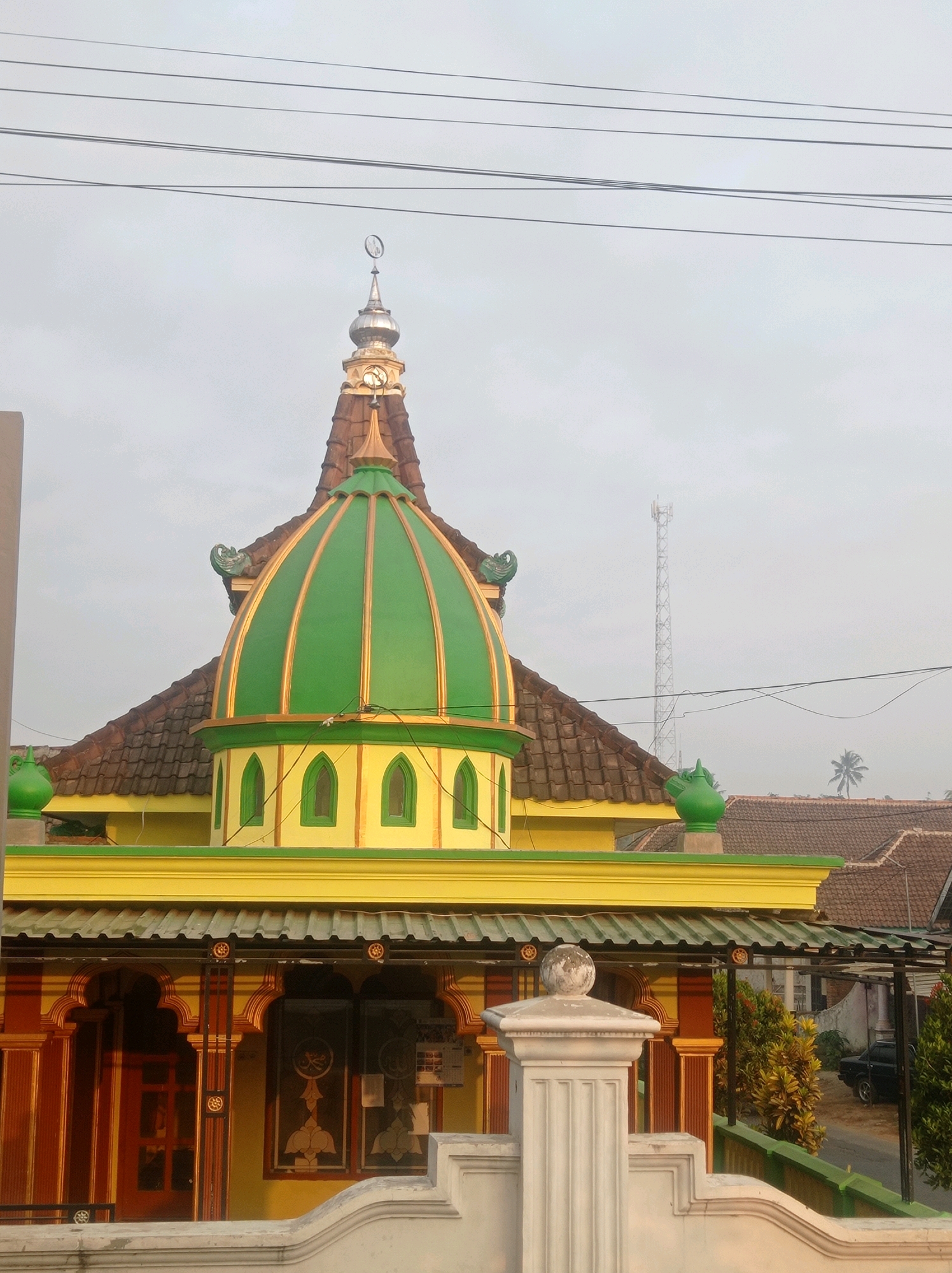 The image shows the exterior of a mosque in Purwosekar, Indonesia. The mosque has a distinctive green dome with gold accents, situated on a yellow and red building with intricate architectural details. The dome is topped with a silver spire featuring a crescent moon and star symbol. In the background, a cell tower and a palm tree are visible against a hazy sky. The foreground features a white concrete wall and railing, with glimpses of trees and a black car. The scene appears to be captured during daylight hours, possibly early morning or late afternoon, given the soft lighting and muted colors.