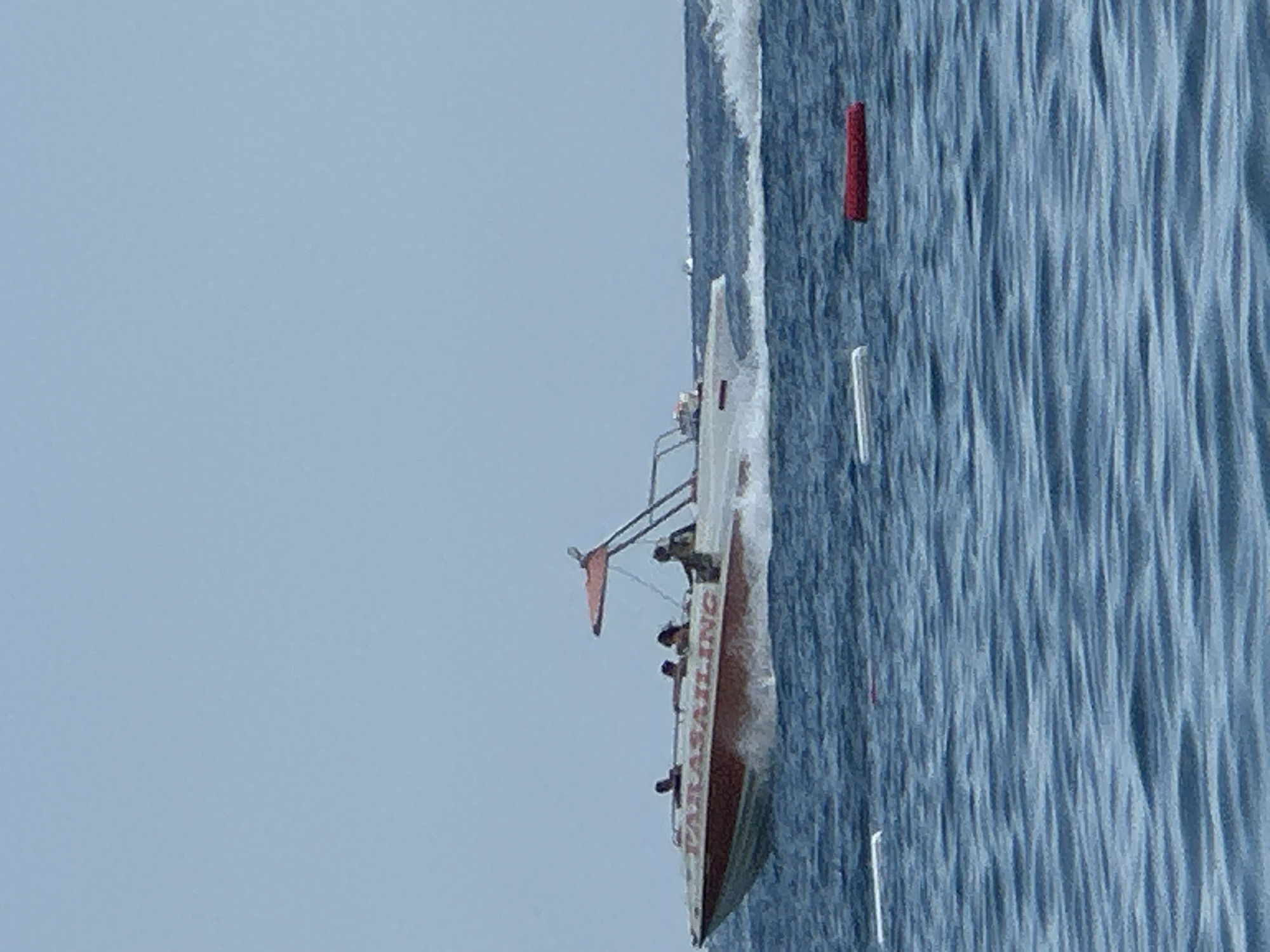 In the city of Boreti, Montenegro, a parasailing boat is seen on the water. The boat is white with red lettering that reads "PARASAILING." Several people are on the boat, some appearing to be crew members and others passengers. The boat is moving, creating a wake behind it. The water is blue and choppy, with several white and red buoys visible in the foreground. The sky is a pale blue and overcast. In the background, other distant boats and landmasses are faintly visible.