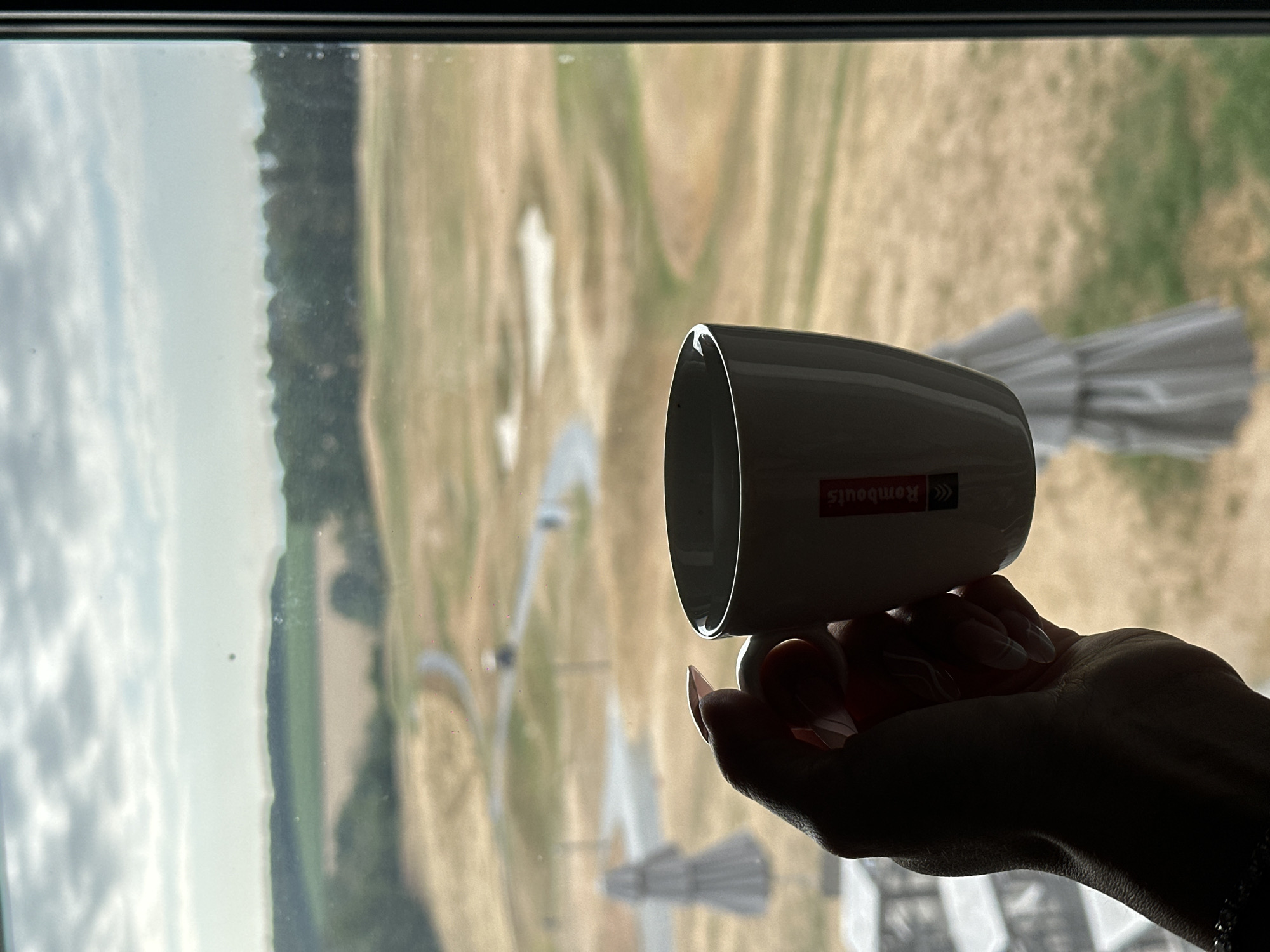 A person's hand, with manicured nails, holds a white ceramic mug with a red logo that says "Rombouts" against a blurred background of a golf course. The background shows green fairways, sand traps, a winding path, and a line of trees in the distance under a cloudy sky. The image is shot through a window, with reflections visible on the glass. The location is Méan, Belgium.