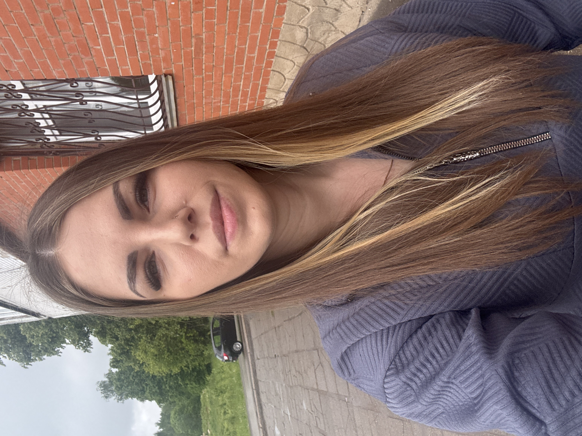 A woman poses for a selfie outdoors. She has long, light brown hair with blonde highlights. She is wearing a dark blue zip-up sweater. A brick building with a barred window is in the background. A car and trees are also visible in the distance. The image was taken in Shevlyakovo, Russia.