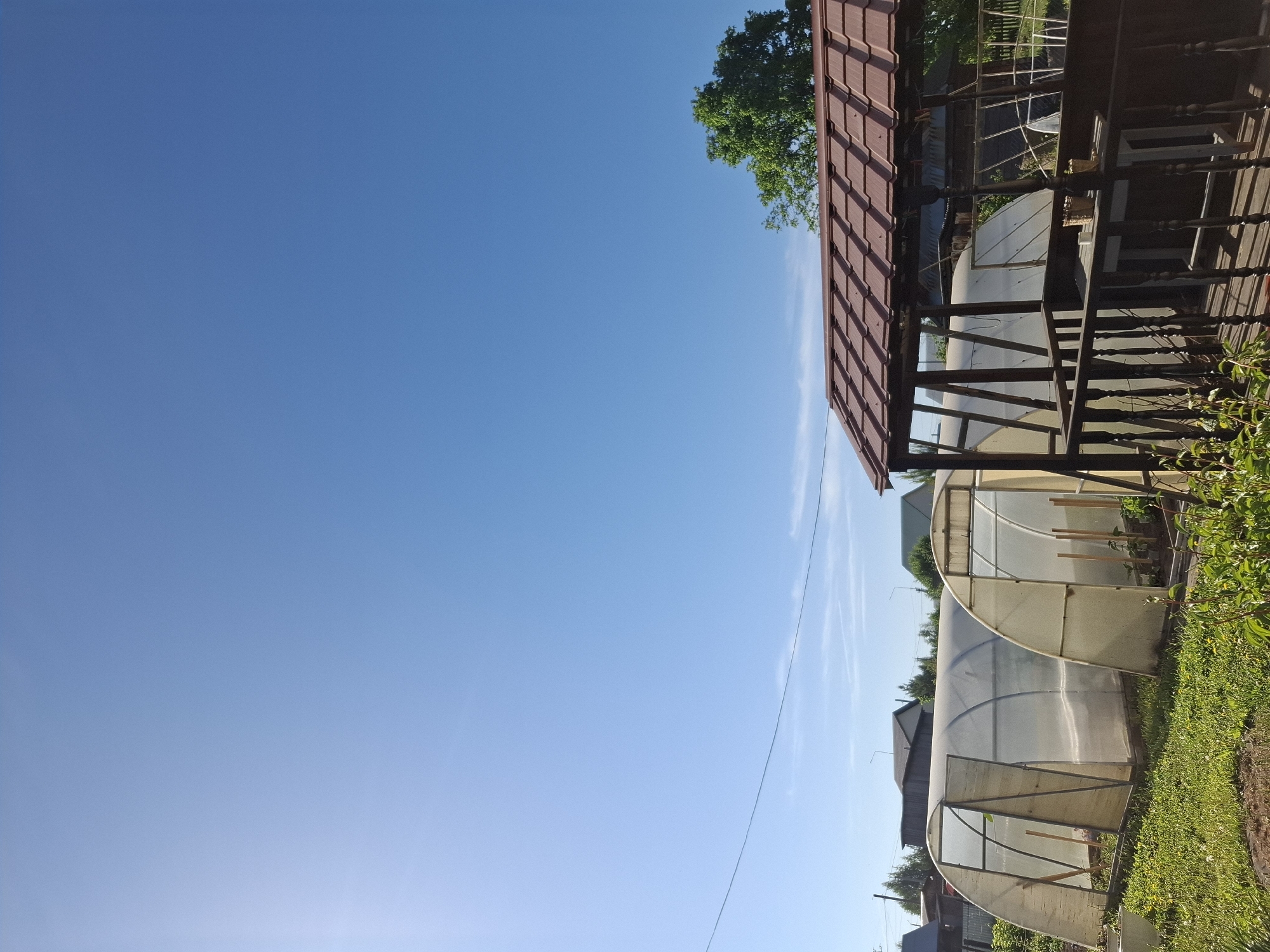 The image depicts a clear blue sky above a rural scene in the city of Амзя, Russia. In the foreground, there are several tunnel greenhouses constructed from metal frames and covered with translucent plastic sheeting, standing on a grassy area. Adjacent to the greenhouses is a brown wooden structure, possibly a porch or shed with a tiled roof. Green foliage is visible along the edge of the image, suggesting vegetation or plants.
