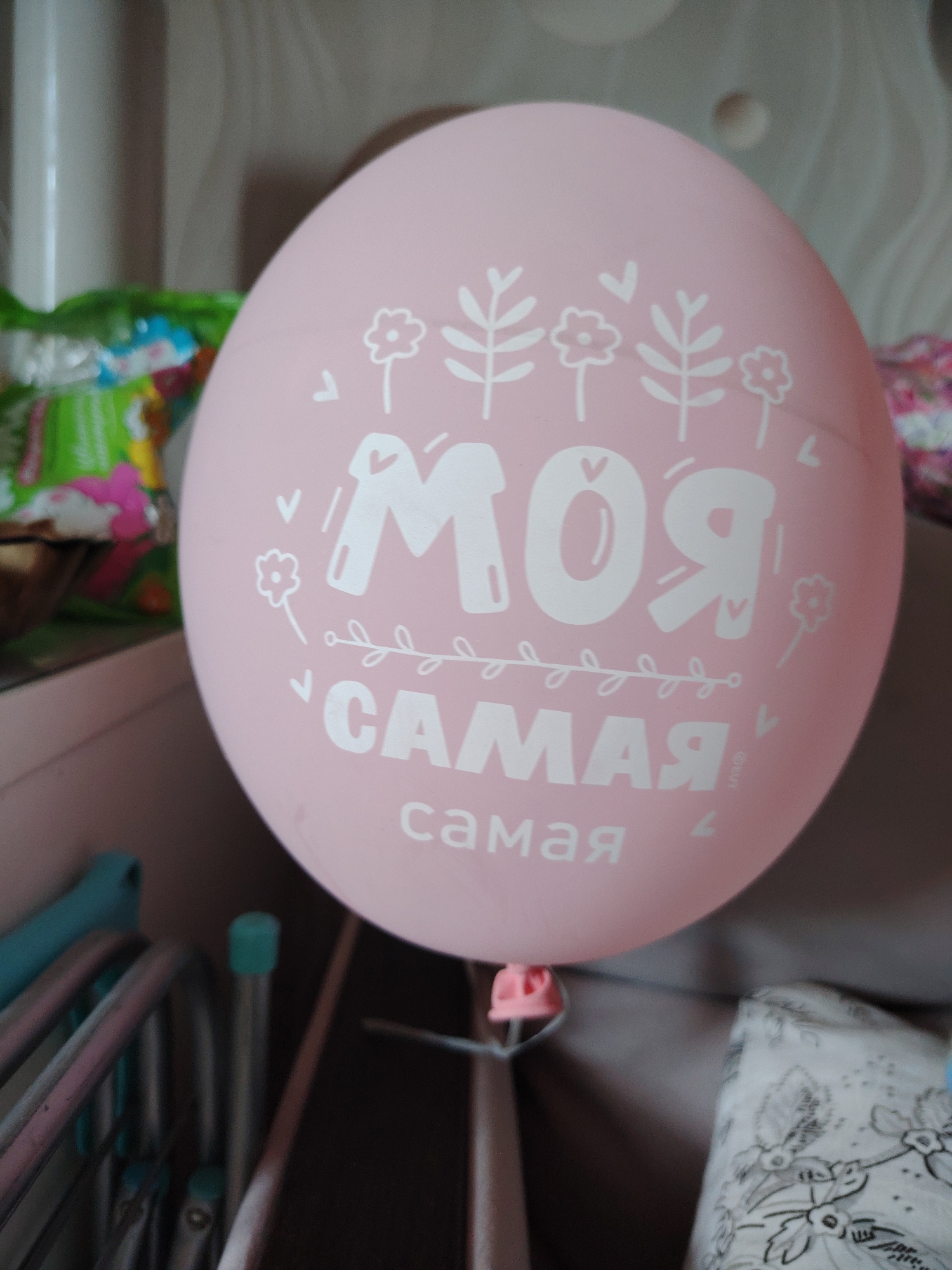 A close-up, slightly out-of-focus image shows a light pink balloon with white text and floral illustrations. The text on the balloon reads "МОЯ САМАЯ" which translates to "MY DEAREST" or "MY MOST". The floral illustrations include stylized flowers and leaves. The balloon appears to be floating or suspended by a thin string. In the background, there are glimpses of other objects, including what appears to be a metal folding chair, a plastic blue cap on a thin tube, and a patterned fabric. The overall environment suggests an indoor setting, possibly a room in a residential building in the city of Khimki, Russia. The lighting is soft, indicating it may be daytime with indirect light.