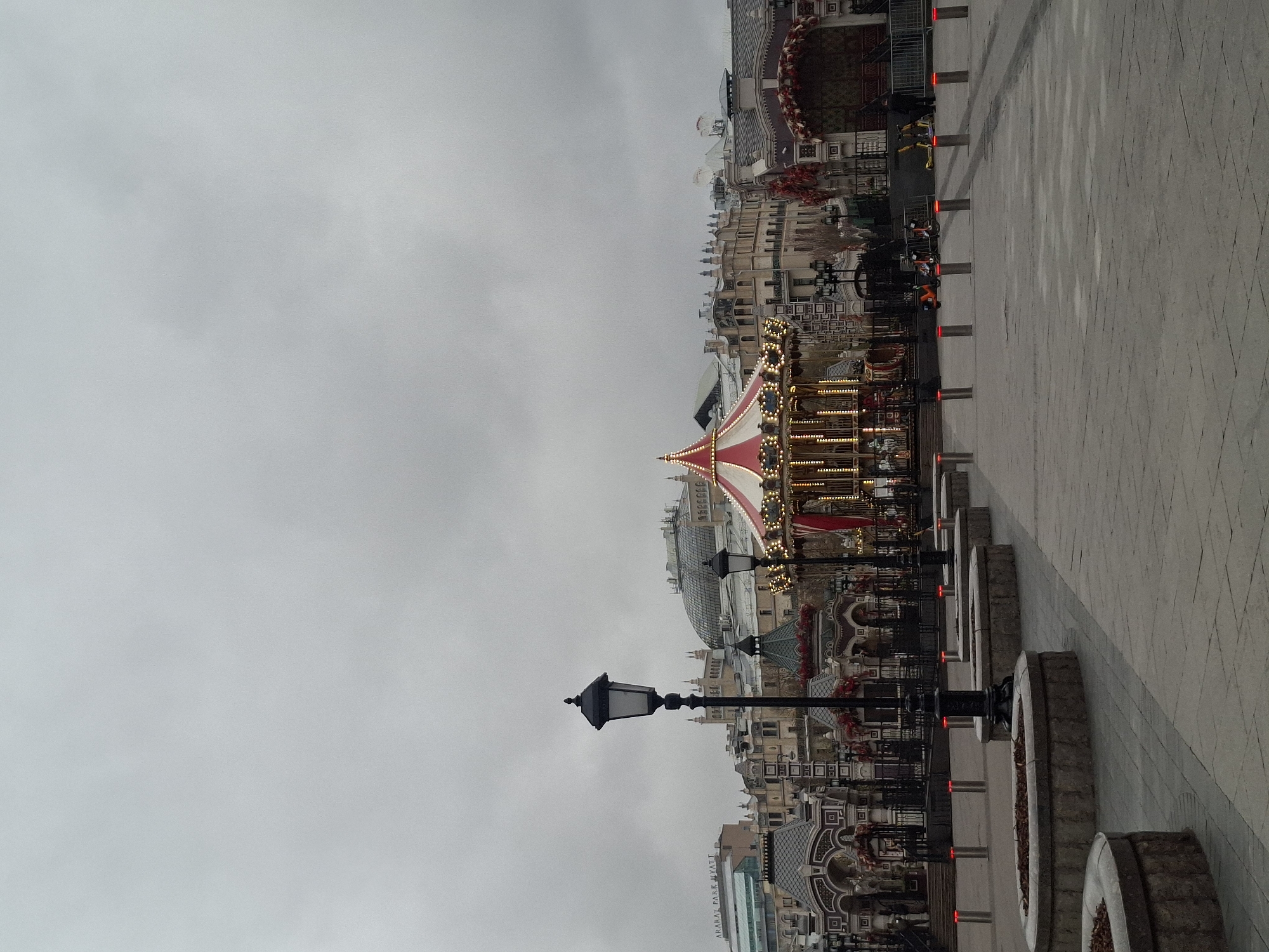 This media file captures a wide shot of a public square or promenade on an overcast day. The image is framed to show a blend of architectural detail and festive elements under a uniformly grey sky.
The central feature is a large, classic-style carousel positioned on the left side of the frame, adorned with a distinctive red and white striped canopy and numerous golden, warm lights.
A paved walkway, patterned with light-colored stones, stretches from the foreground towards the background on the right. This walkway is bordered by a series of dark bollards, each emitting a red glow from its top. Interspersed along the walkway are ornate, black lampposts, with one prominent in the near foreground. Stone planters or decorative elements are also visible along the path.
In the background, behind the carousel and lampposts, stands a complex of multi-story buildings. These structures feature intricate architectural details, including ornate facades, many windows, and some distinctive domed roofs. Portions of the buildings, particularly their upper sections, appear to be decorated with festive arrangements, possibly wreaths or garlands. On one of these background buildings, the text "ARARAT PARK HYATT" is clearly visible.
The absence of people in the immediate vicinity contributes to a quiet atmosphere. The overcast sky suggests a gloomy day, or potentially dusk, with the illuminated carousel and bollard lights indicating a low light setting. The scene depicts a grand urban space, likely a commercial or entertainment area, consistent with a major city like Moscow, Russia.
