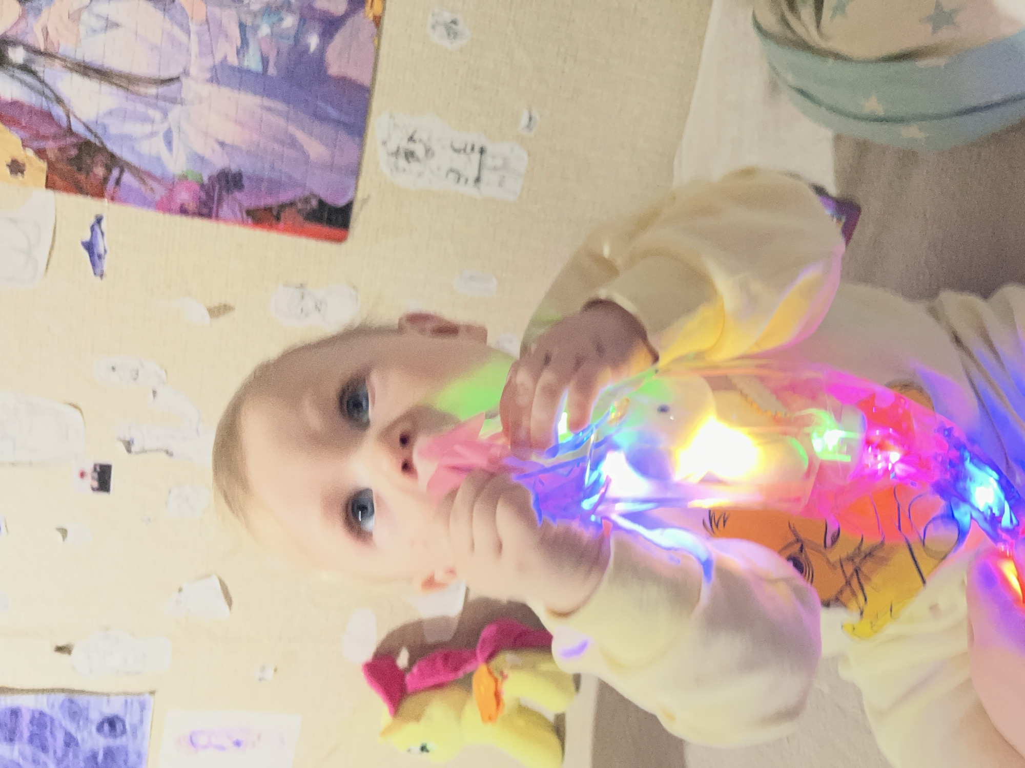 A baby with blond hair and blue eyes is holding a plastic light-up toy. The baby is wearing a yellow shirt with an image of Winnie the Pooh. A plush My Little Pony toy is near the baby's hand. In the background, there is a light beige wall adorned with stickers and a purple artwork. A pillow with blue stars is visible in the upper right corner. The location and city details are not discernable from the image.