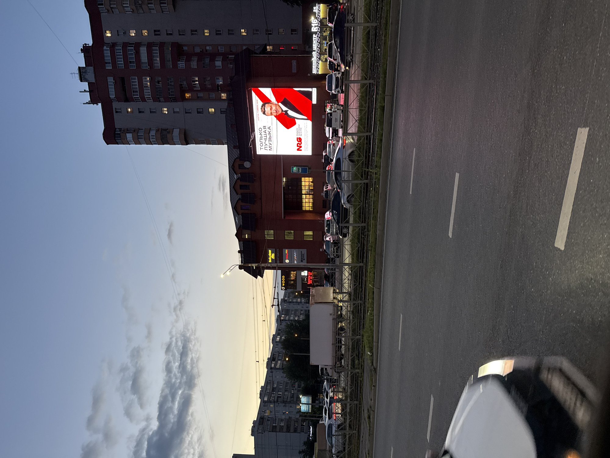 The image depicts a street scene in Kazan, Russia, during twilight. A large, illuminated billboard dominates the upper right portion of the frame, displaying an advertisement for "NRG" radio. The billboard features a portrait of Justin Timberlake with the text "ТОЛЬКО ЛУЧШАЯ МУЗЫКА" (Only the best music) and "РАДИО СТАНЦИИ" (Radio station) along with "92.3 FM".
In the background, multi-story apartment buildings are visible, some with lit windows, suggesting residential occupancy. Several cars are on the road, indicating moderate traffic. A white truck is parked on the side of the road. The environment is urban, with streetlights and power lines present. The sky is a mix of dusk colors, with clouds scattered across it. The foreground shows a road with white lane markings and a car in the lower left corner.